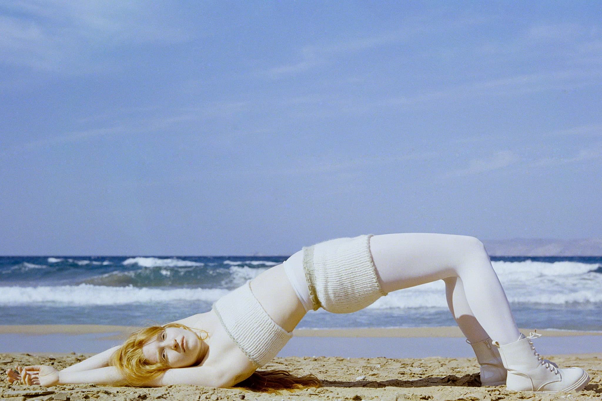 Person in white outfit posing on a sandy beach with ocean waves in the background.