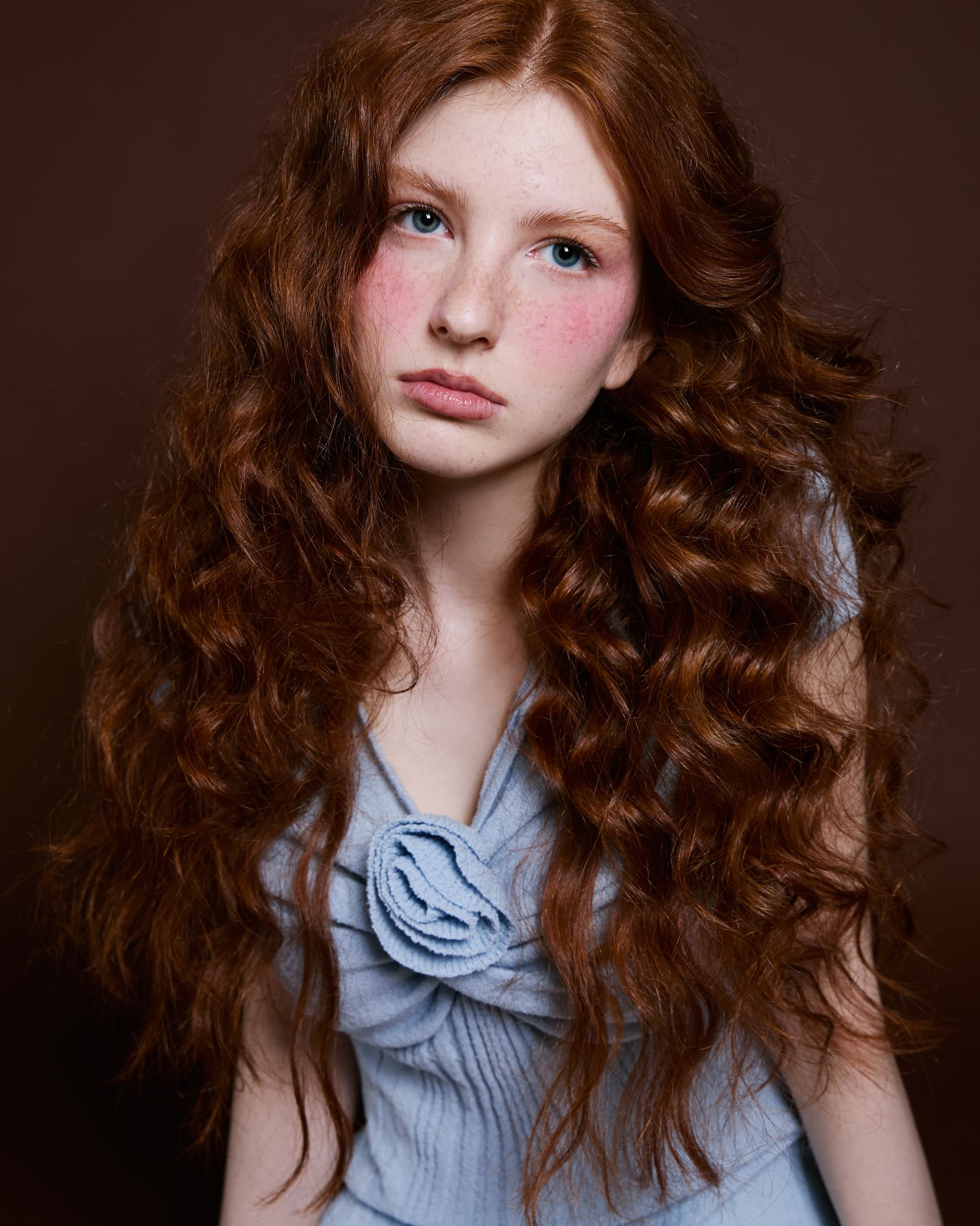 Young woman with long curly red hair and a blue top against a brown background.