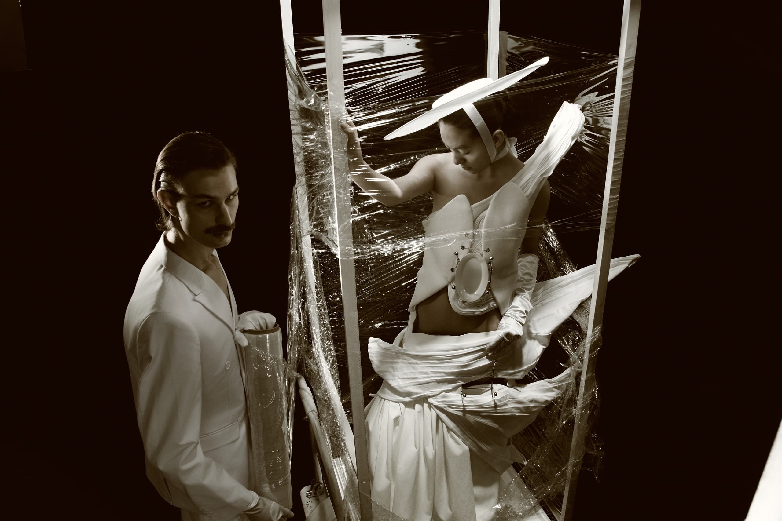 Avant-garde fashion art with figures wrapped in cellophane, dramatic lighting.