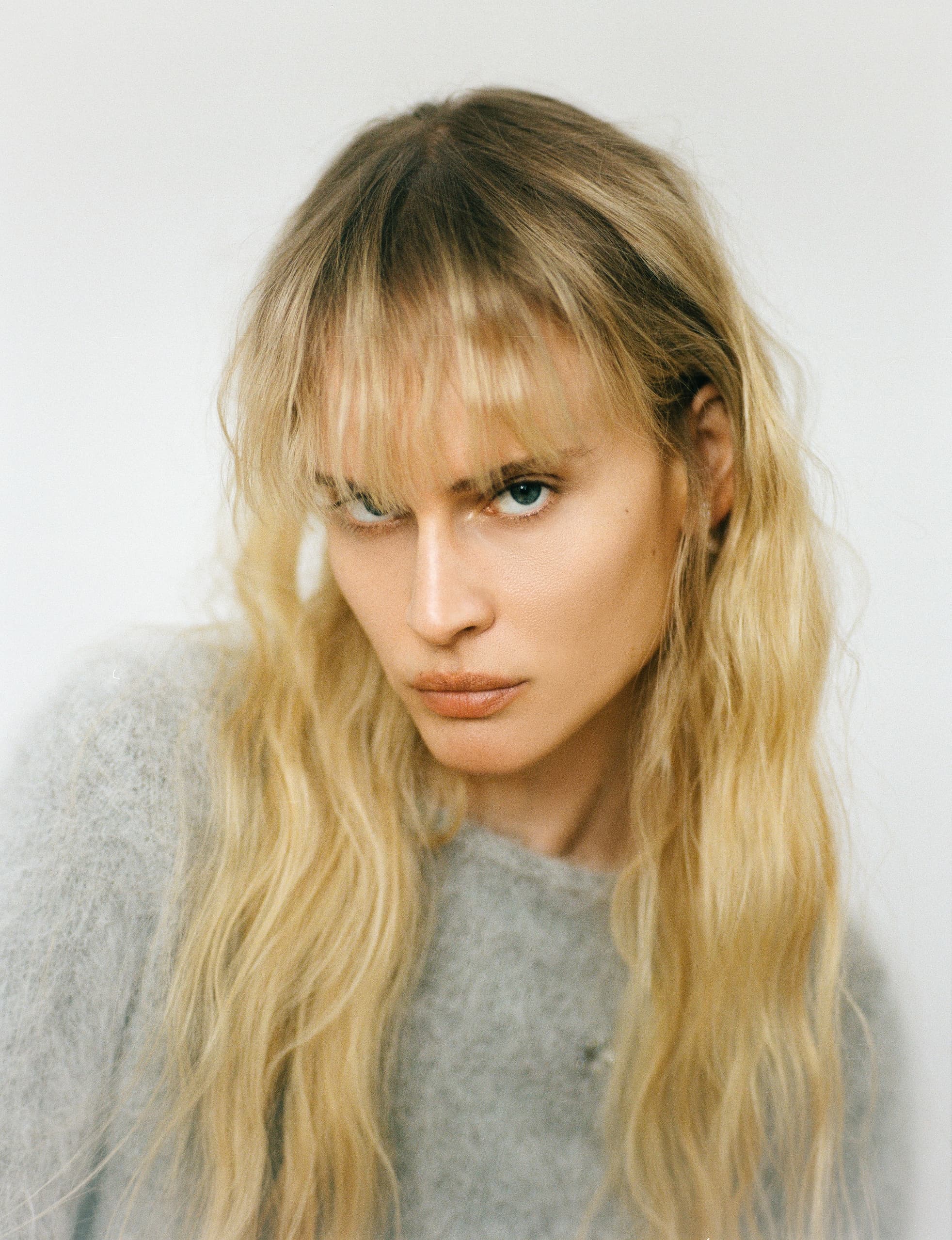 Blonde woman with soft waves and bangs wearing a gray sweater, gazing intently.