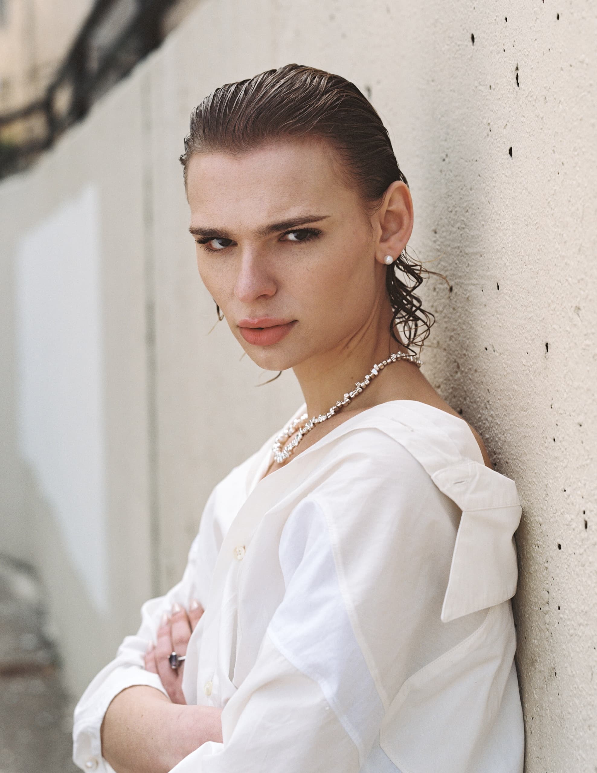 Person with slicked back hair, white shirt and necklace leaning against a wall.