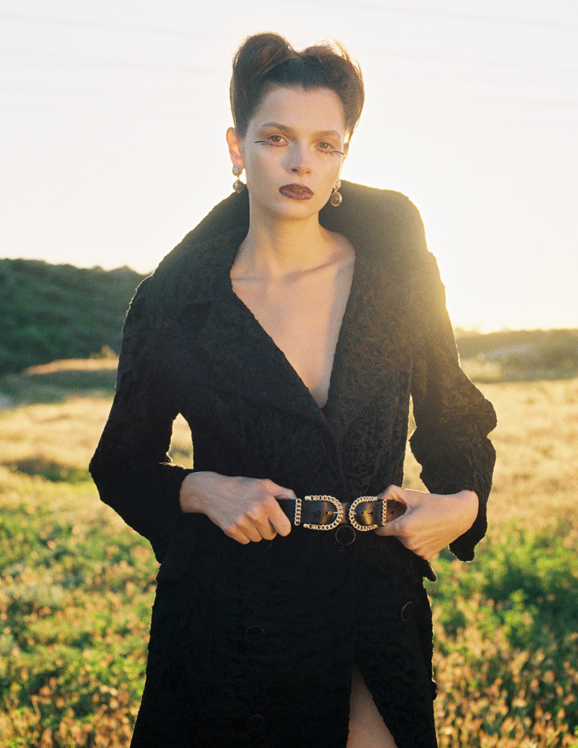 Stylish woman in black coat at sunset, standing in a scenic field.