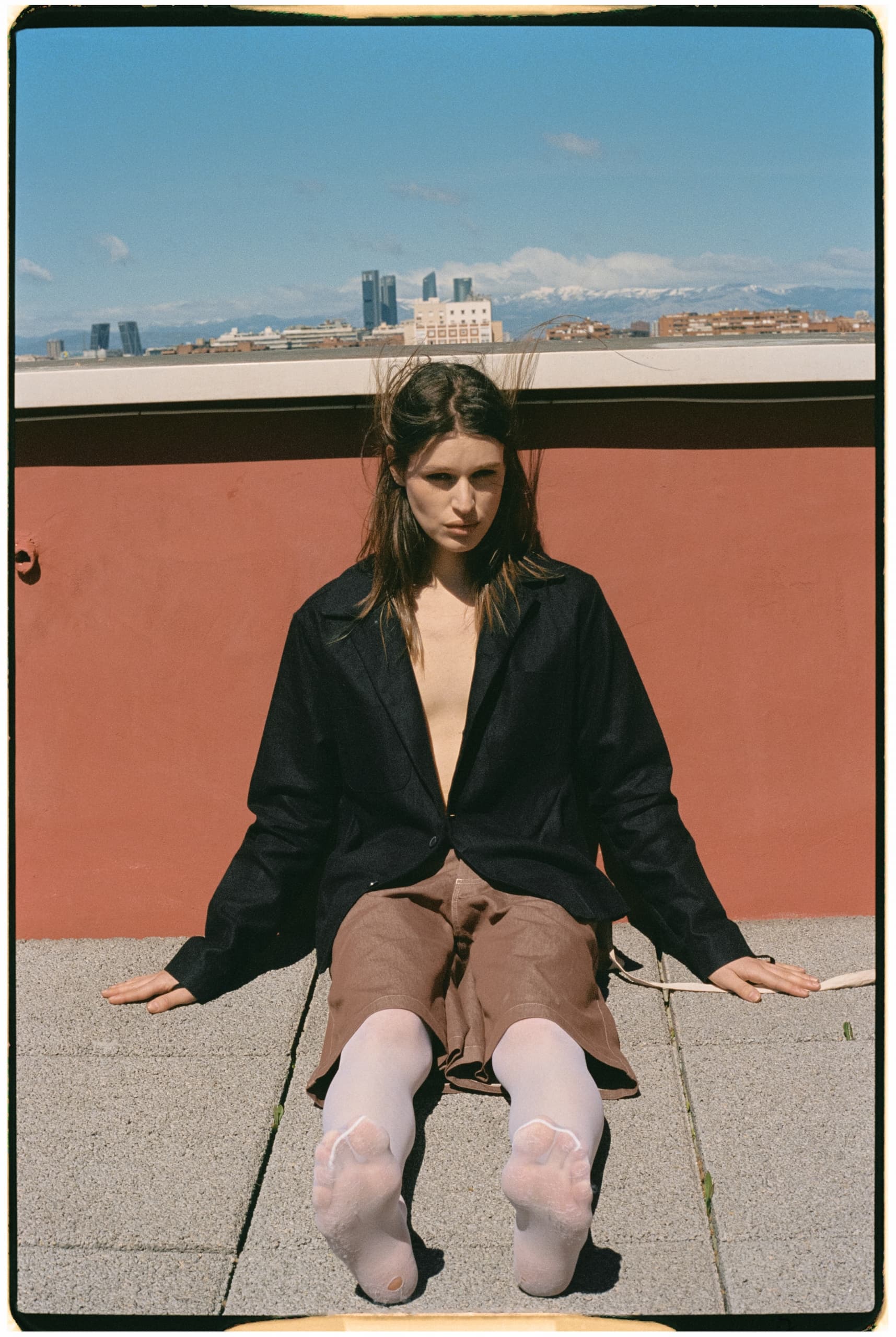 Person sitting on rooftop in city, wearing jacket and shorts, with urban skyline view.