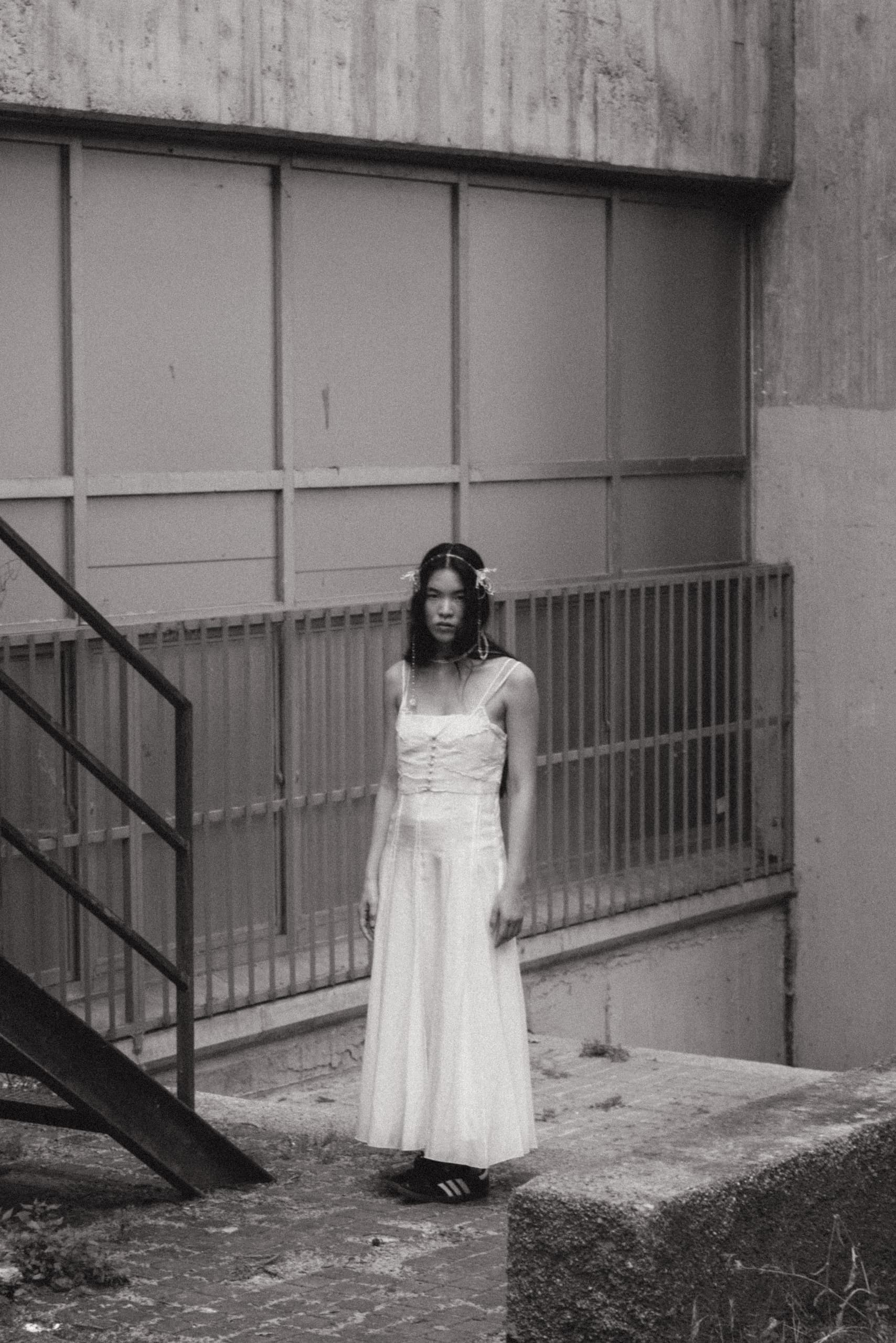 Woman in a white dress standing by stairs, black and white urban setting.
