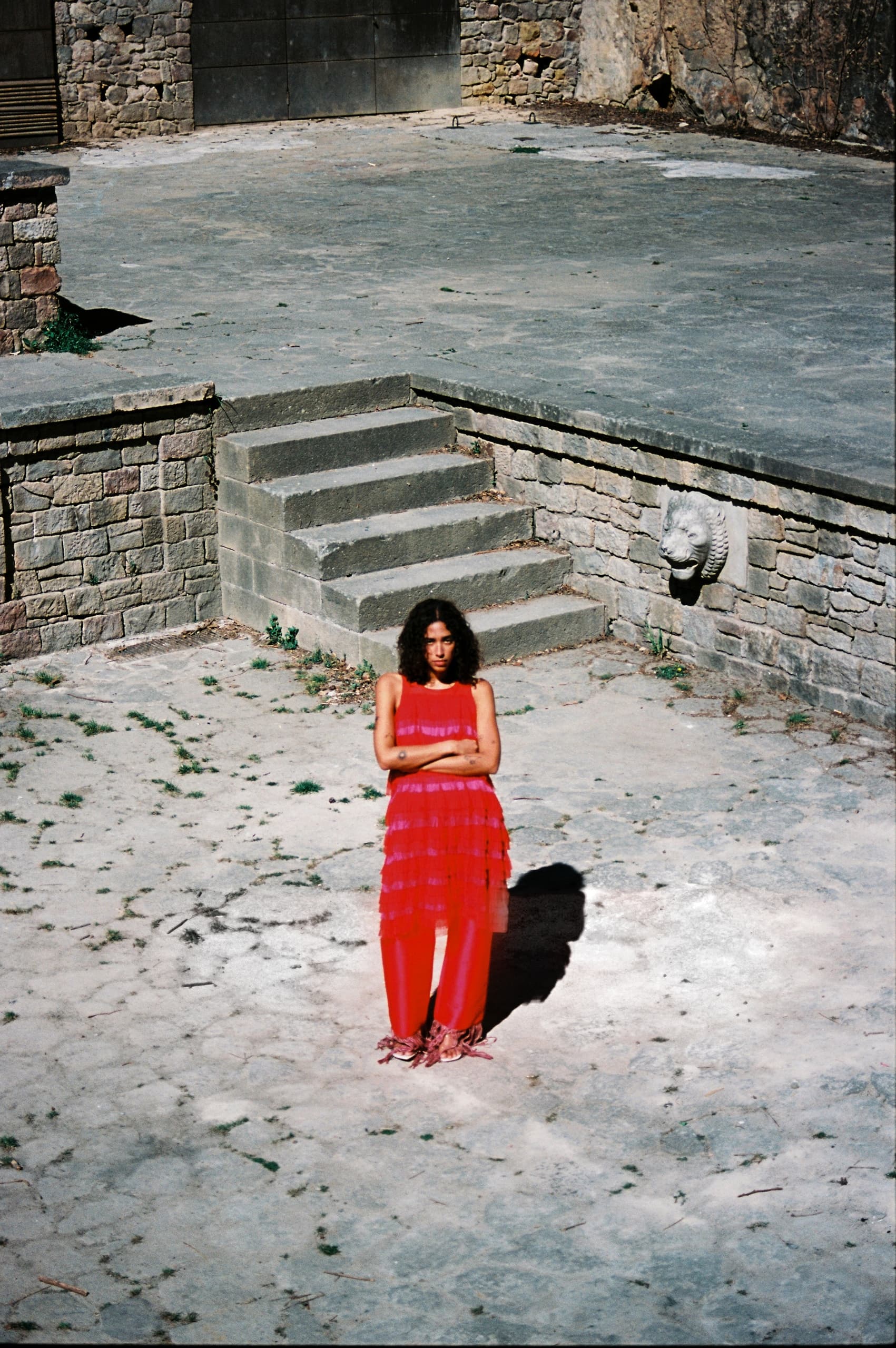 Woman in a red dress standing on a stone courtyard with shadow and stone wall.