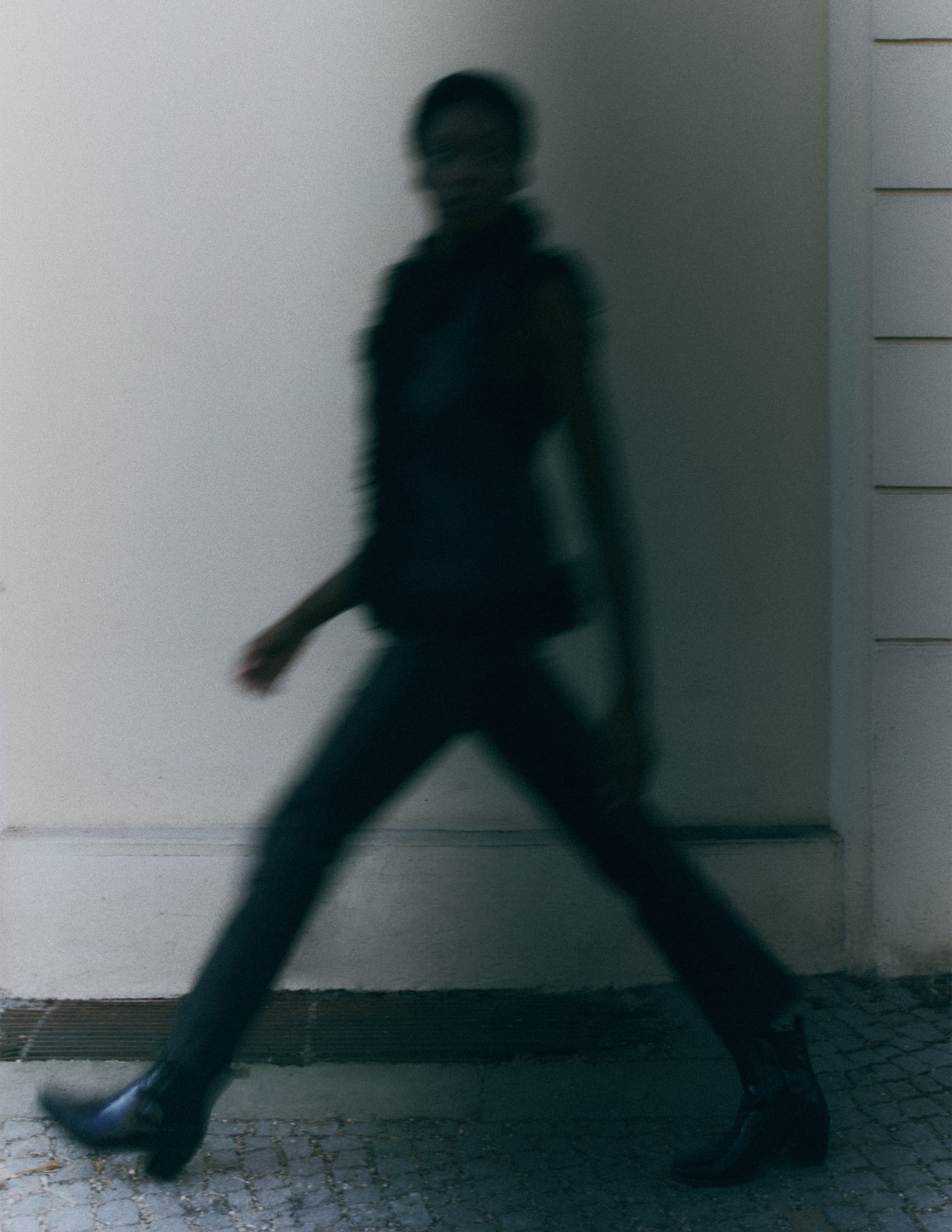 Blurred person walking briskly against a neutral wall background.