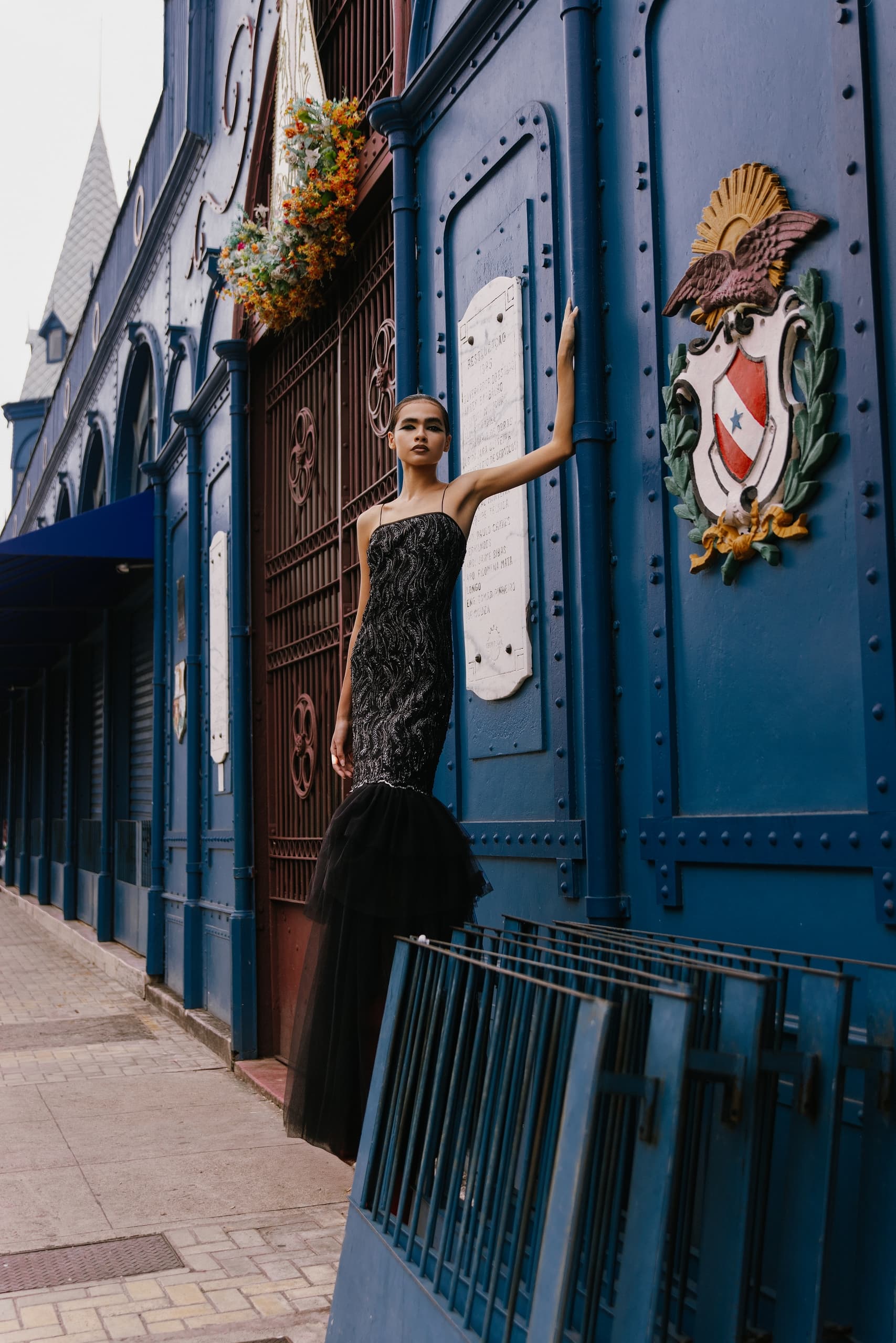 Woman in elegant black dress leaning against ornate blue building.