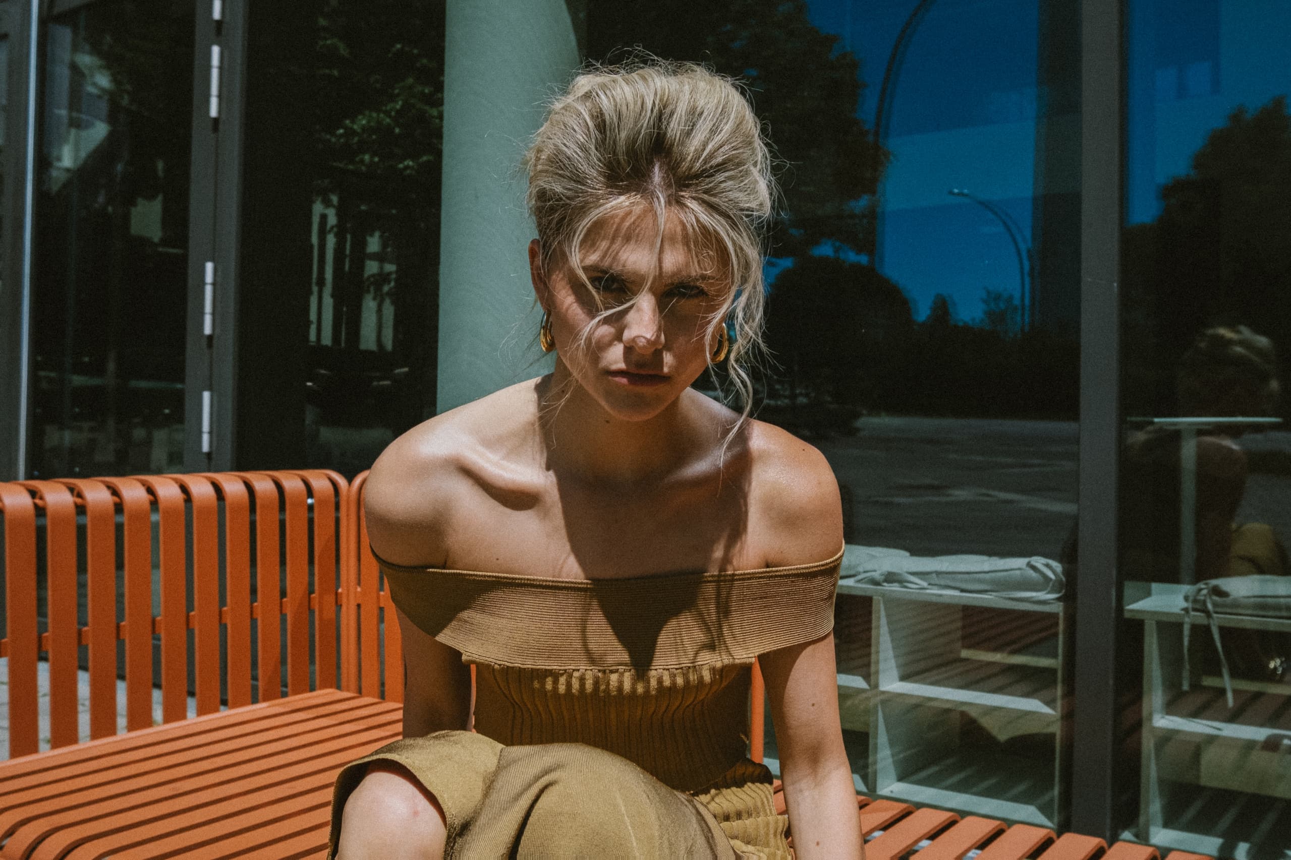 Fashionable woman in off-shoulder dress sitting on orange bench in sunlight.