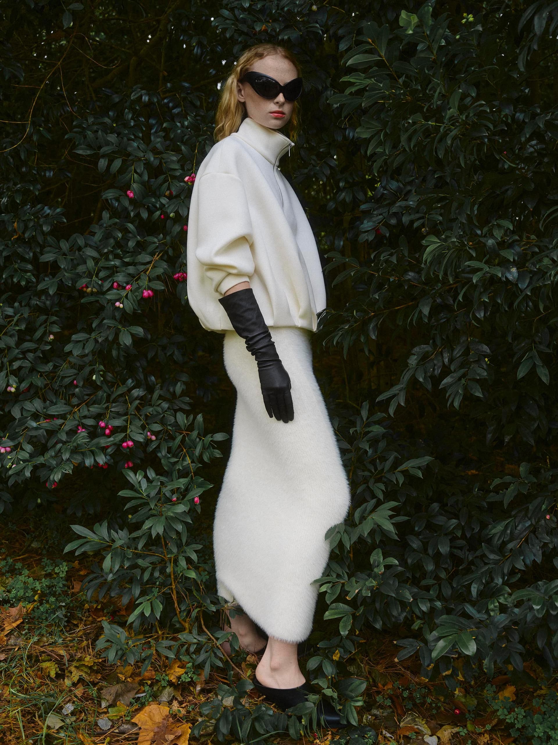 Chic fashion model in white outfit and sunglasses standing amidst lush greenery.