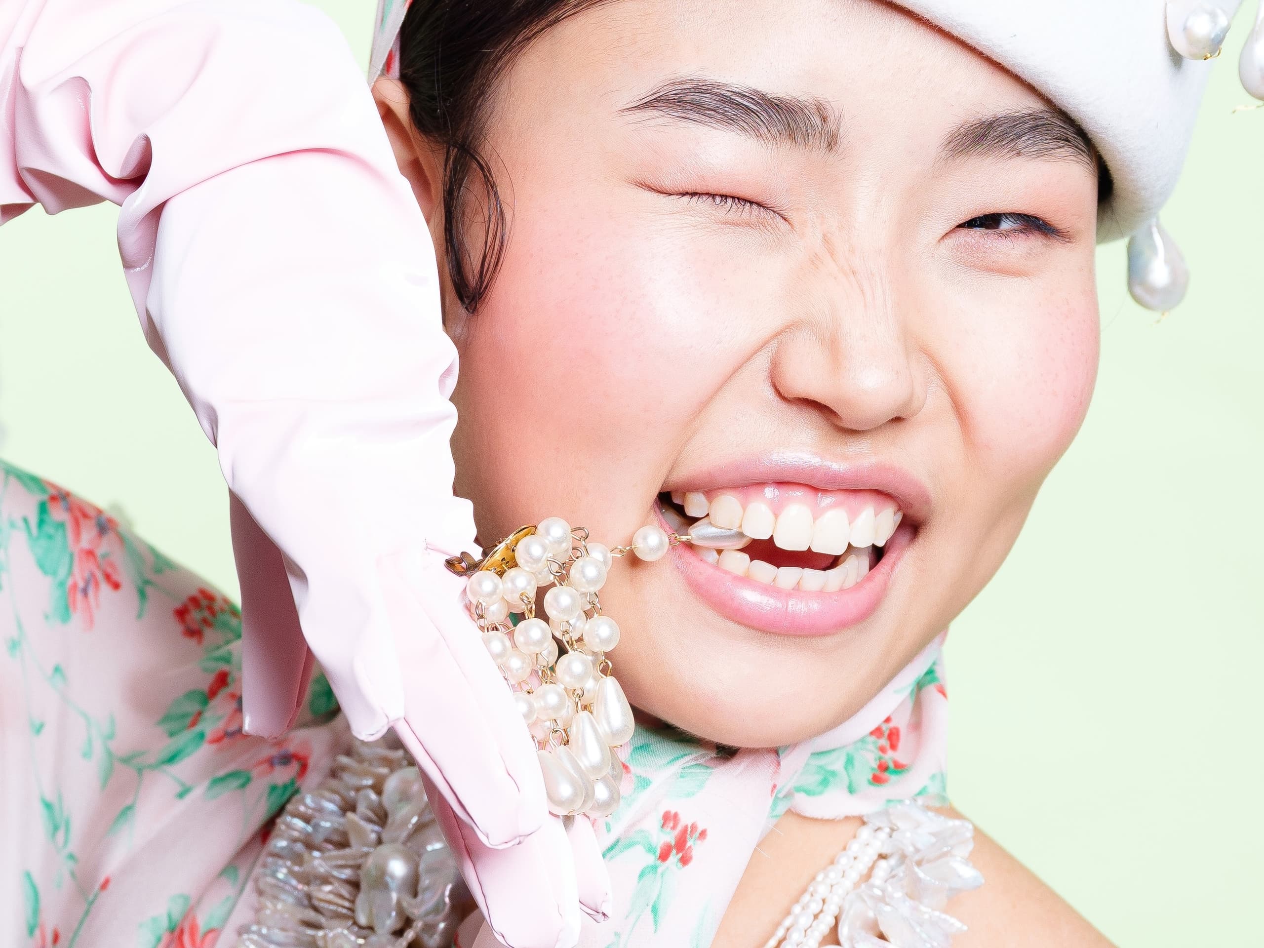 Woman winking, biting pearl necklace, wearing pink gloves and hat, playful fashion.