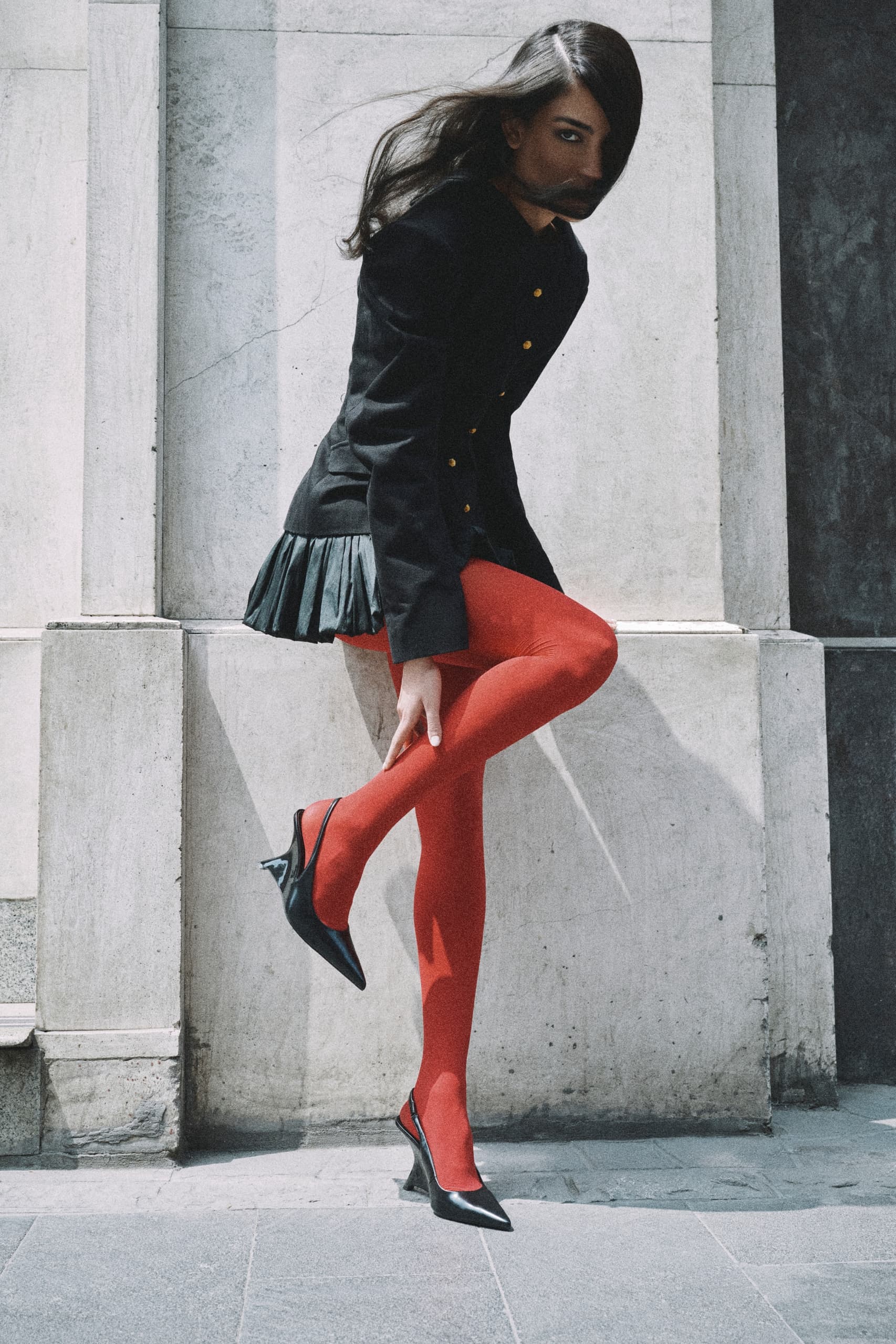 Fashionable woman wearing red tights and black coat poses in urban setting.