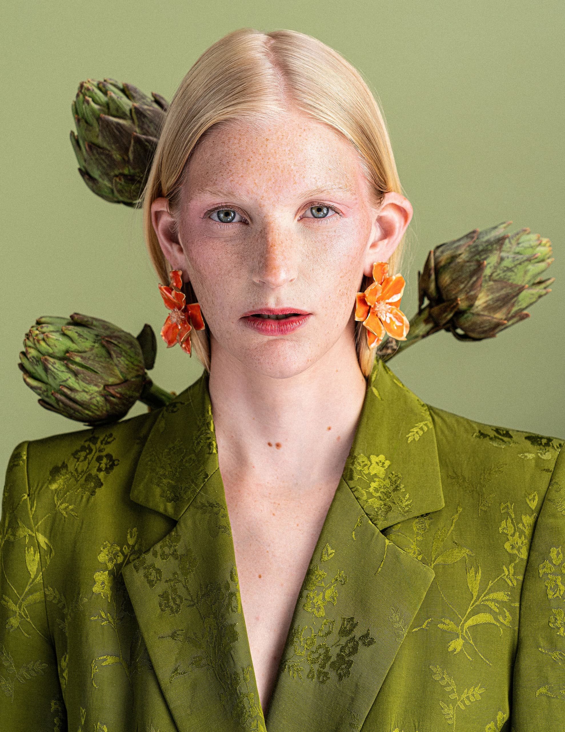 Person in green suit with floral earrings, artichokes in background, green backdrop.