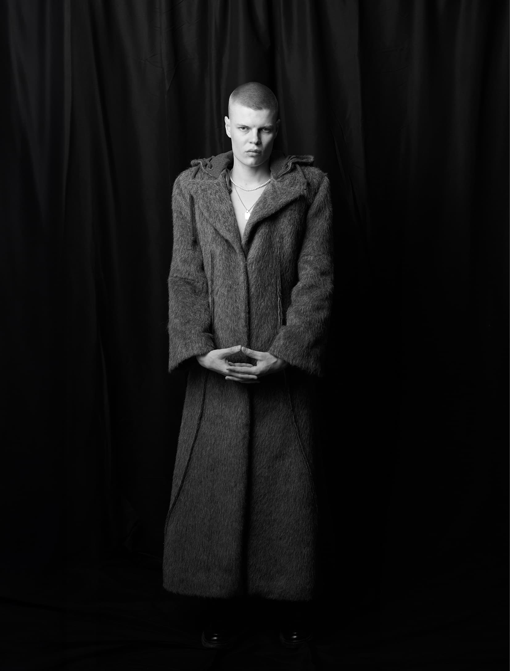 Person in fur coat standing against a dark background in a black-and-white photo.