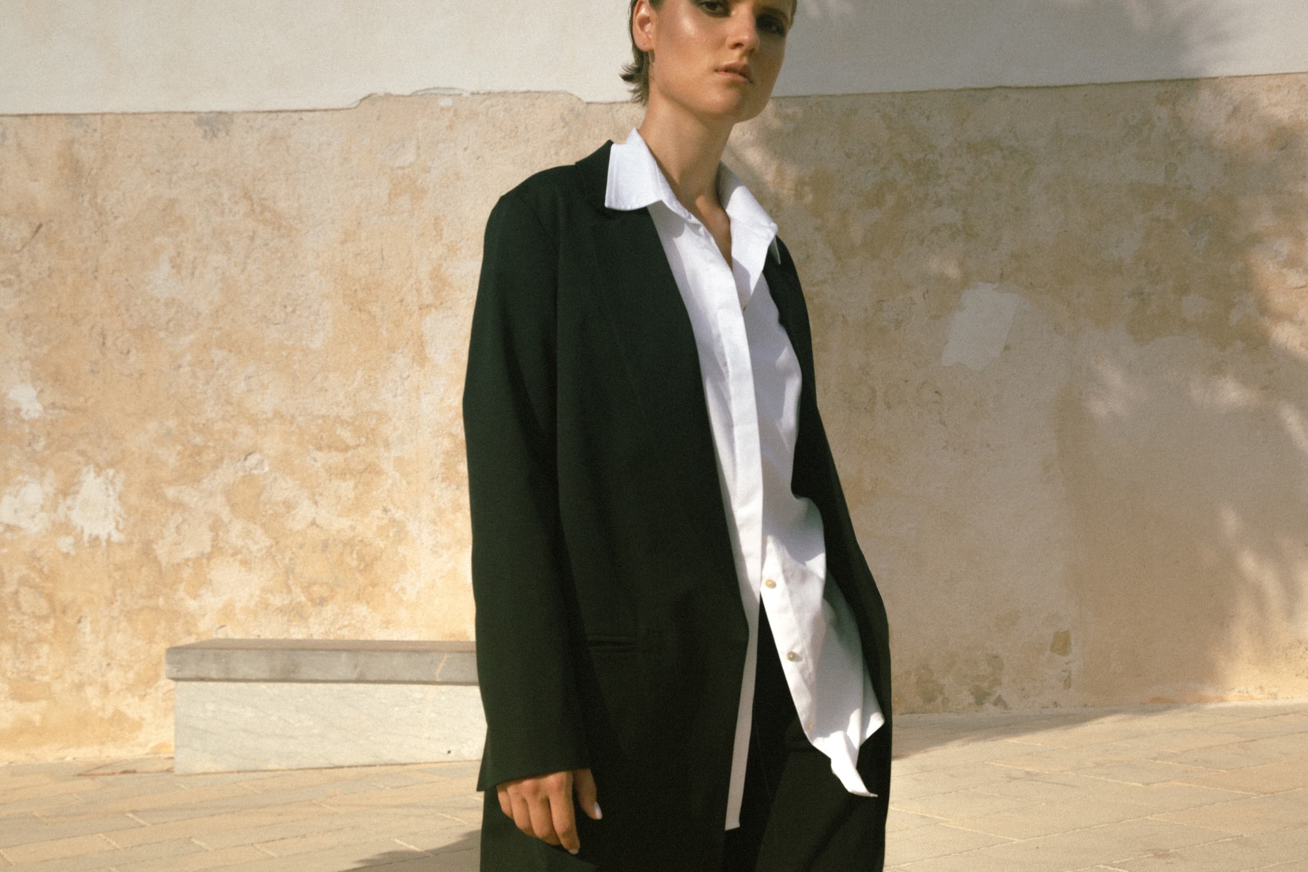 Person in black suit and white shirt standing by a textured wall in sunlight.