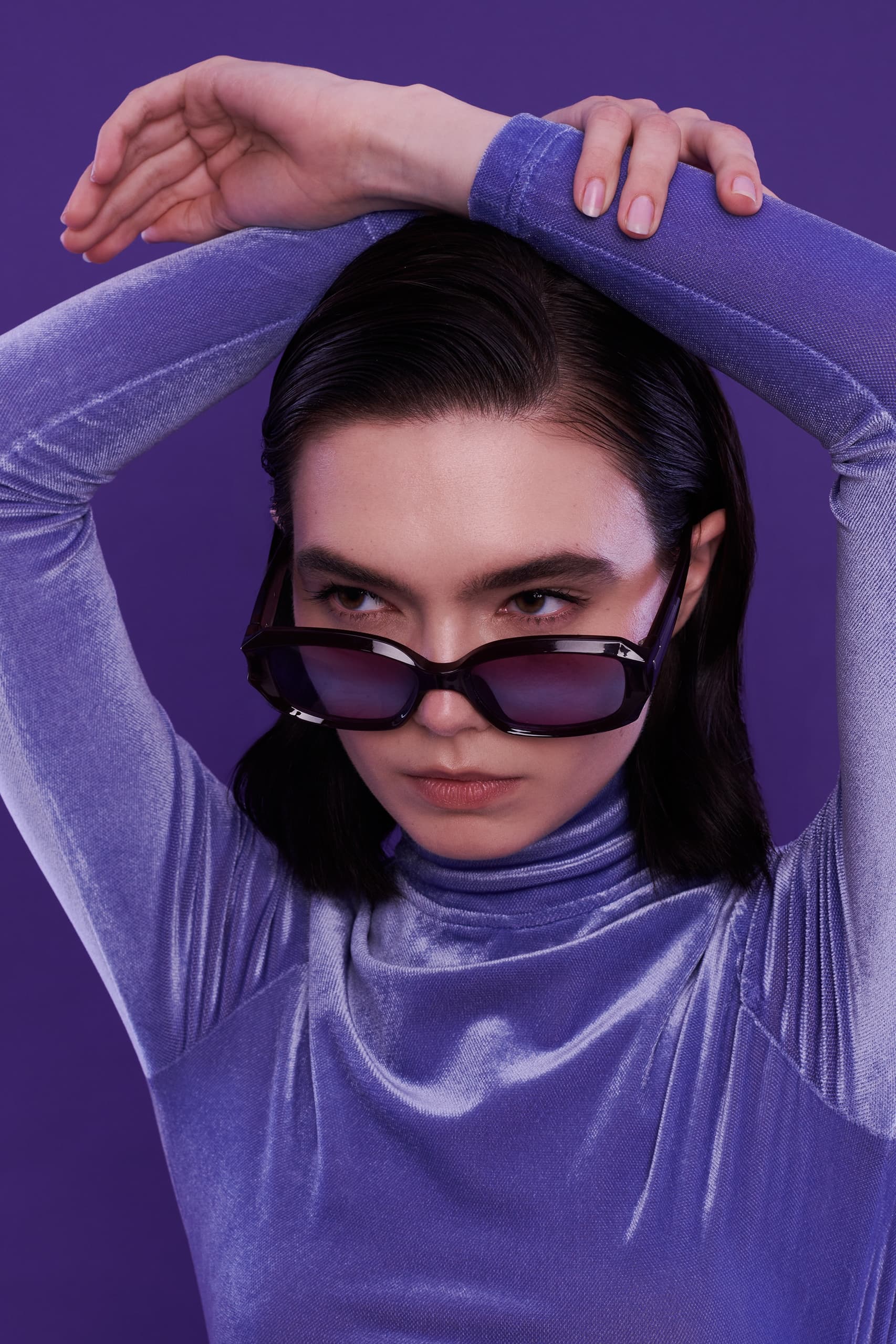 Person in purple velvet top and sunglasses against a purple background.