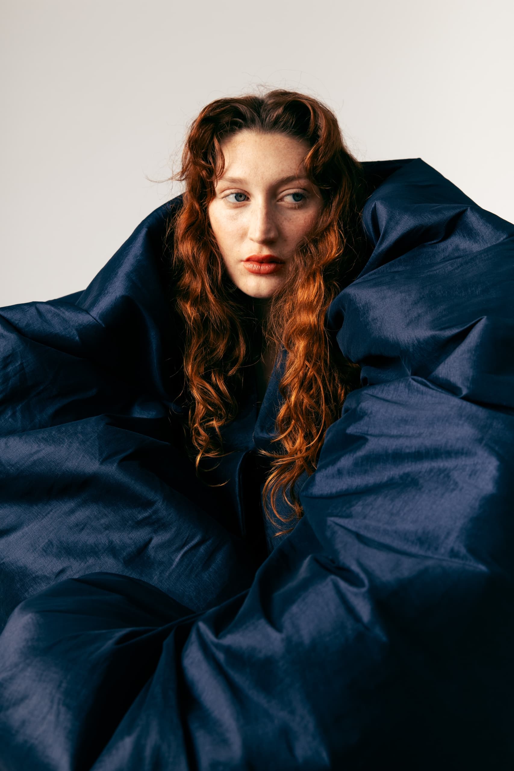 Woman with red hair wrapped in dark blue fabric, gazes thoughtfully to the side.