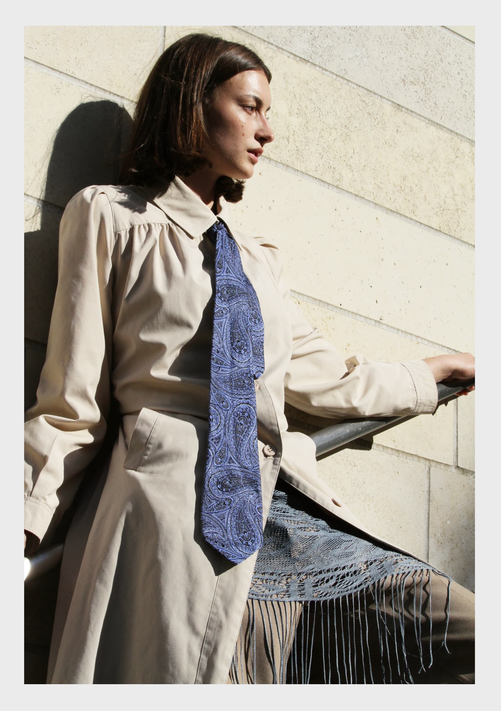 Stylish woman in trench coat and blue tie, posing against a stone wall.