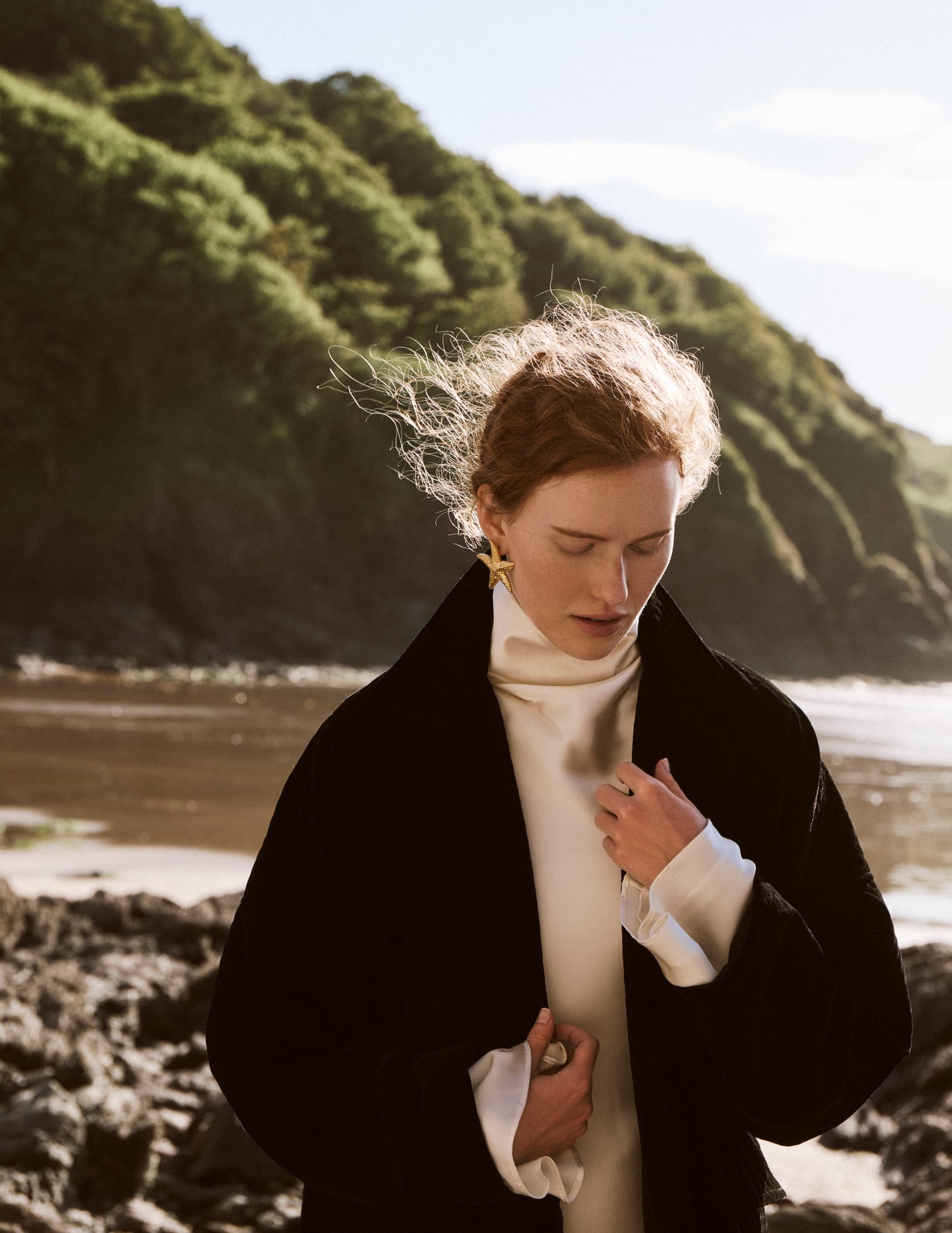 Woman in elegant coat on a rocky beach with green cliffs under a blue sky.