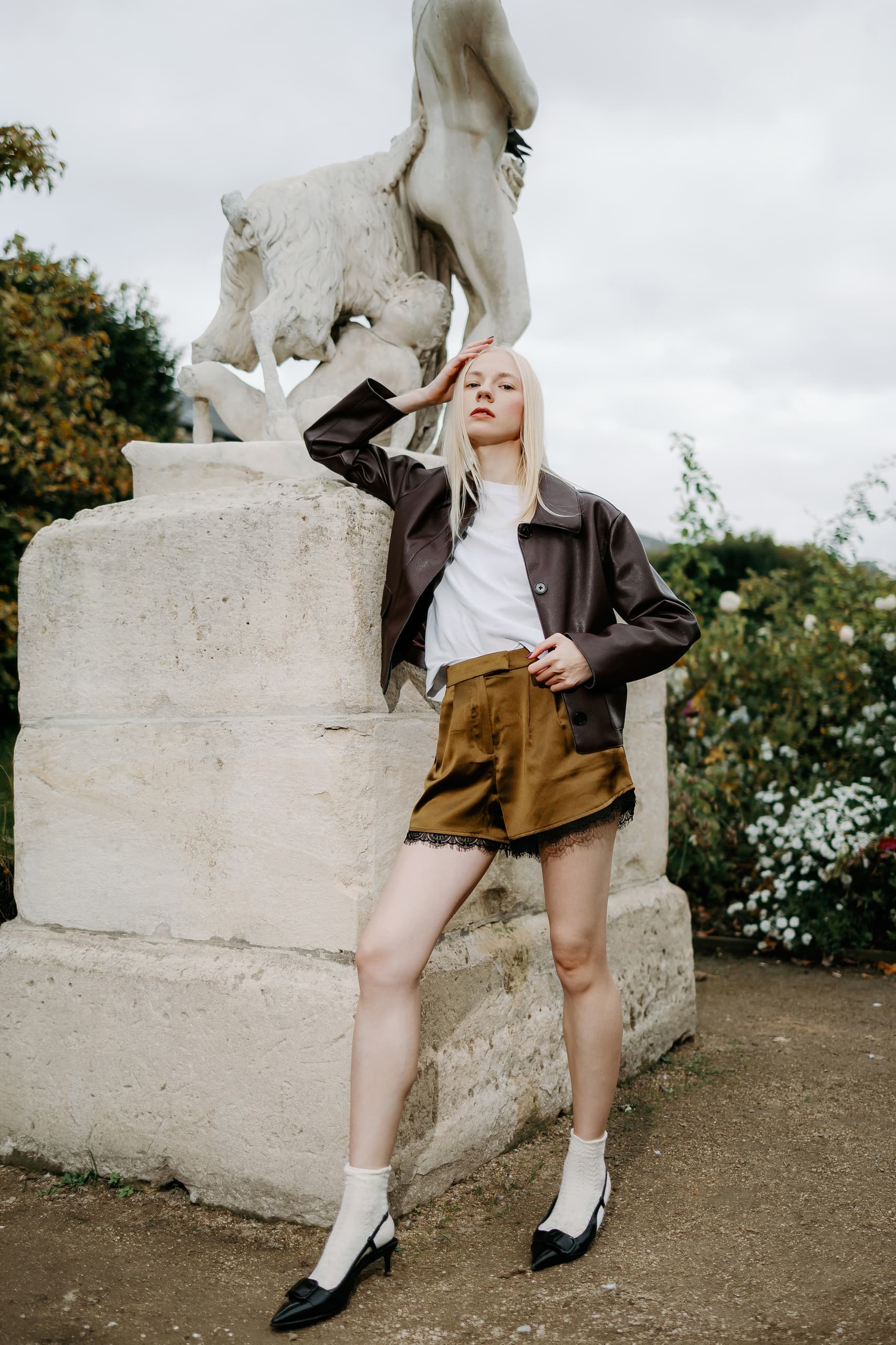 Fashionable woman posing by statue in outdoor garden setting.