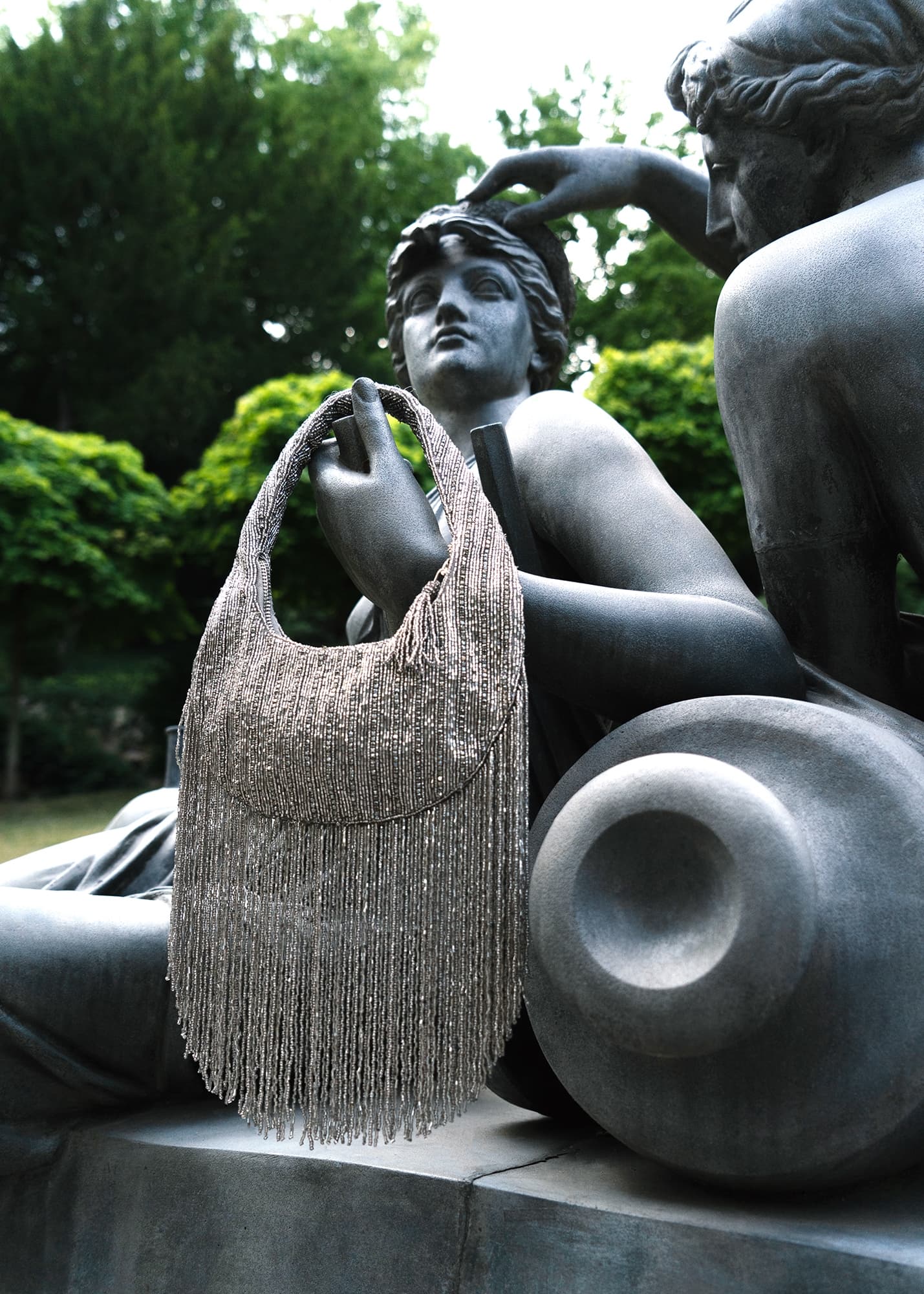 Elegant fringe bag draped on classical statue, surrounded by lush greenery.