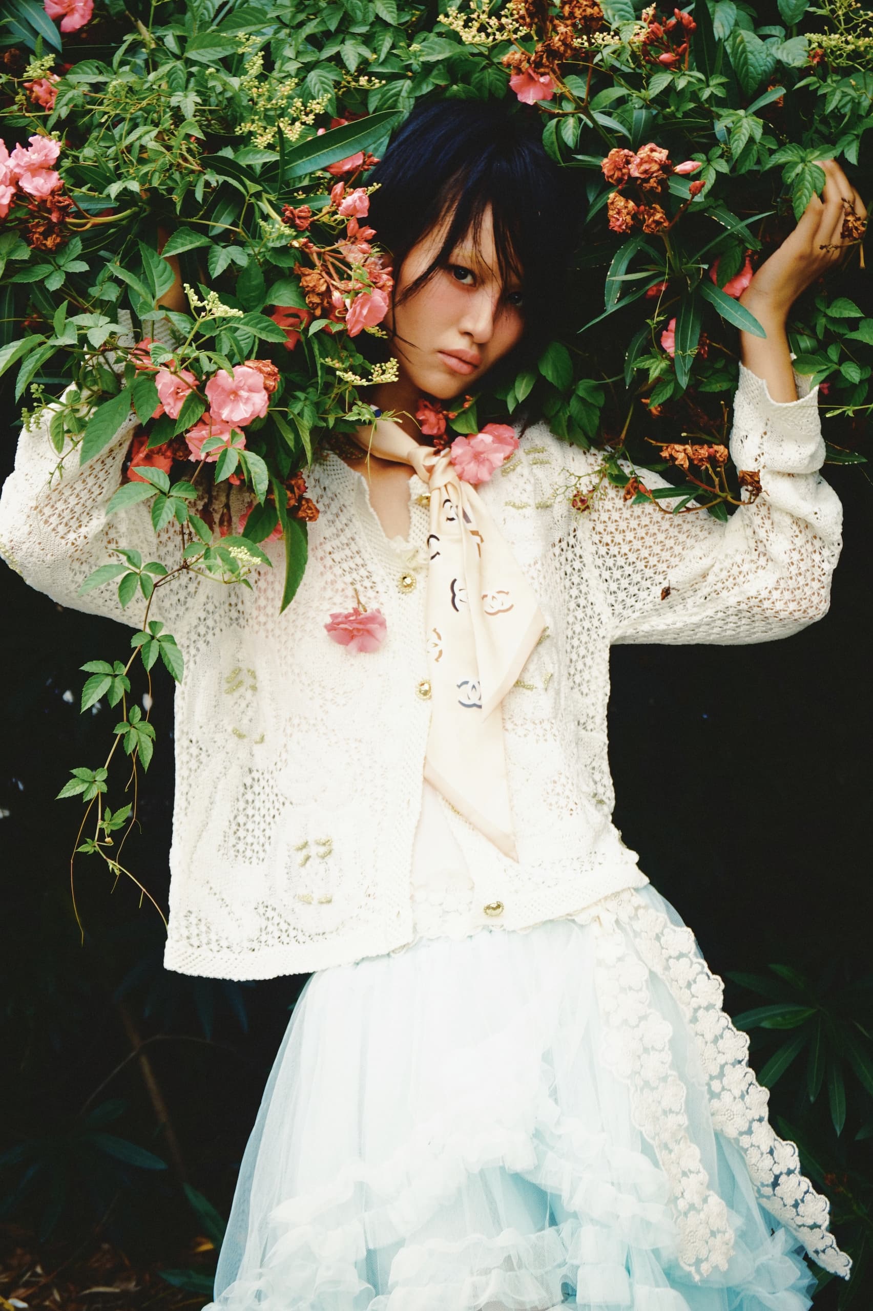 Woman in floral garden, wearing crochet sweater and skirt, surrounded by pink blooms.