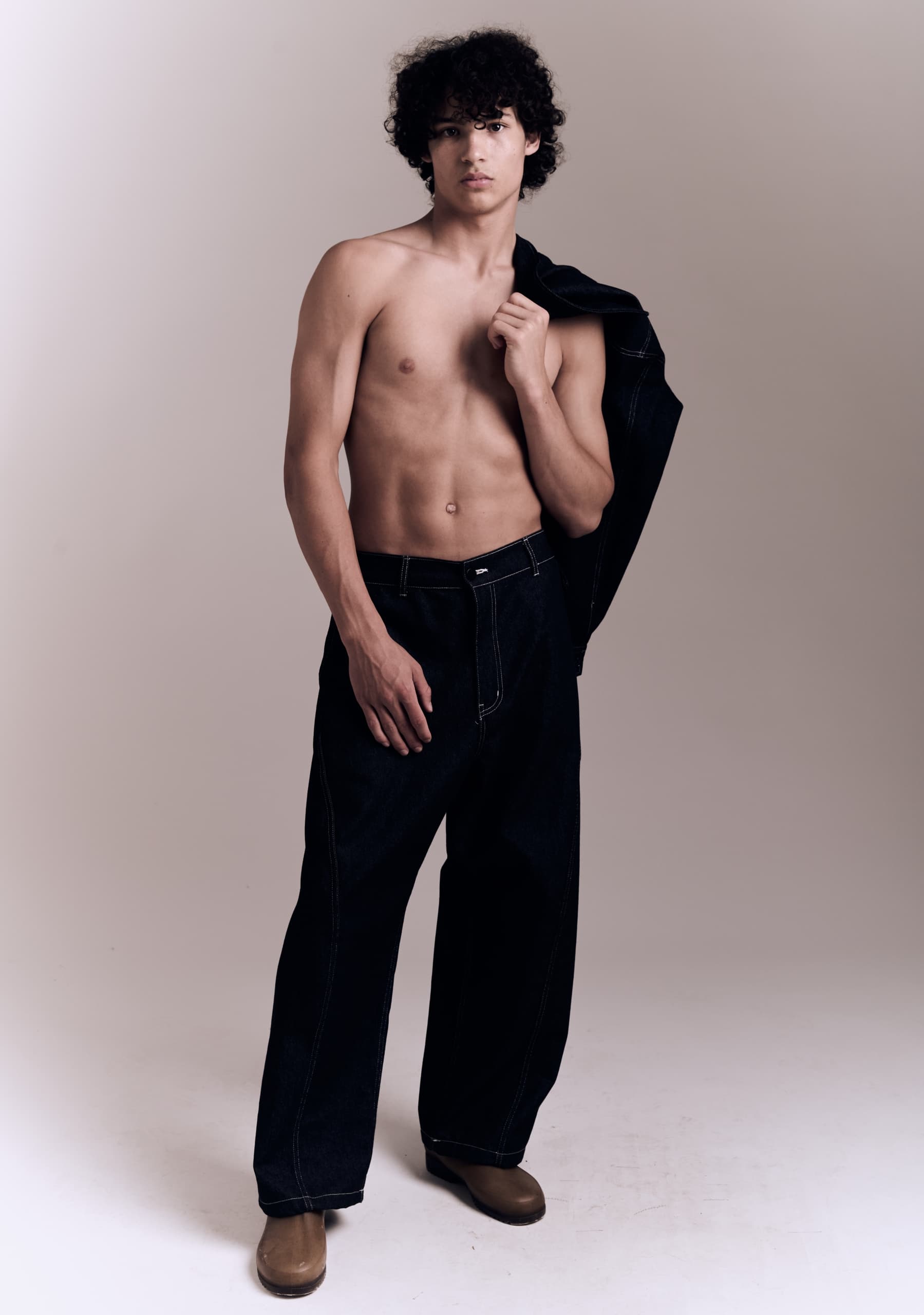Young man posing in dark jeans and boots against beige background.