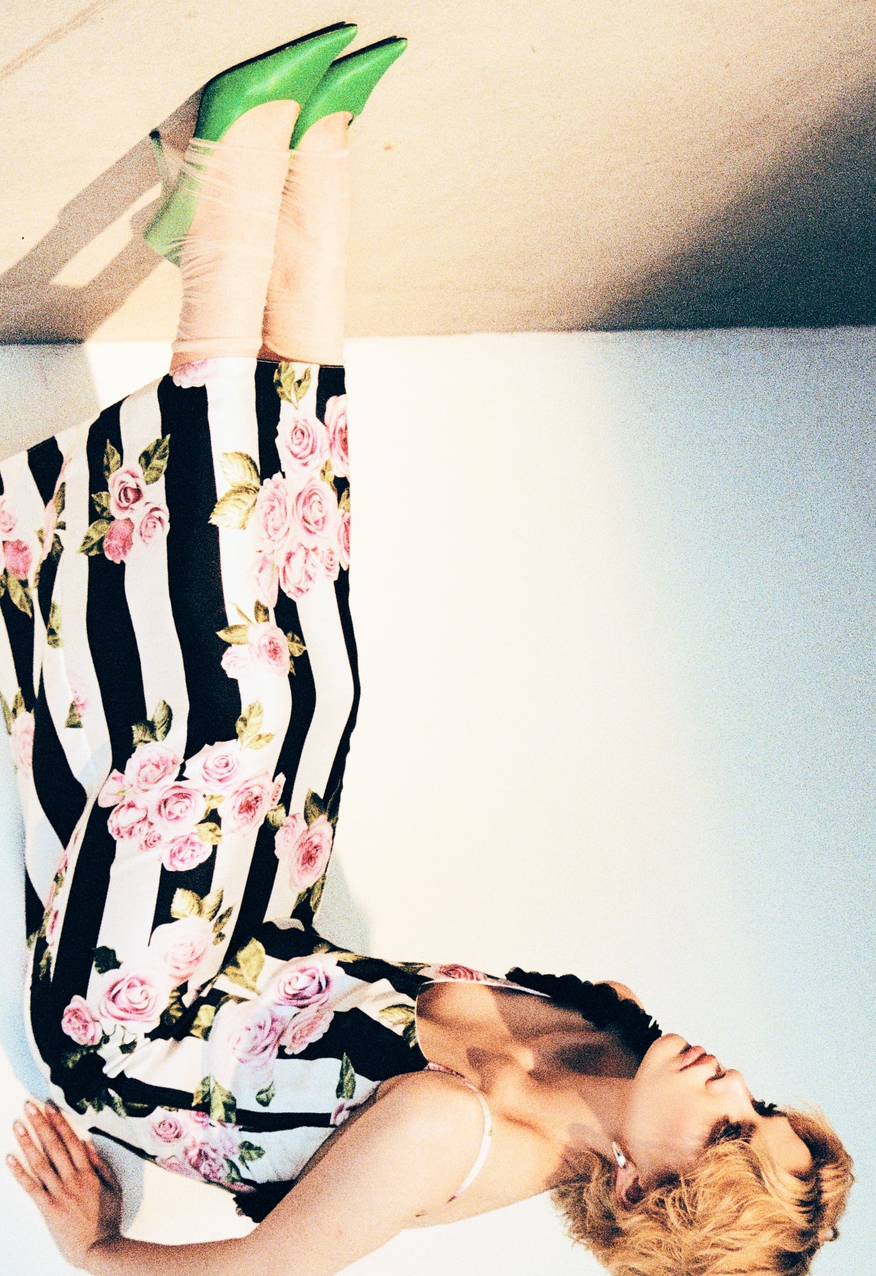 Woman wearing floral striped dress and green heels, leaning on a white wall.