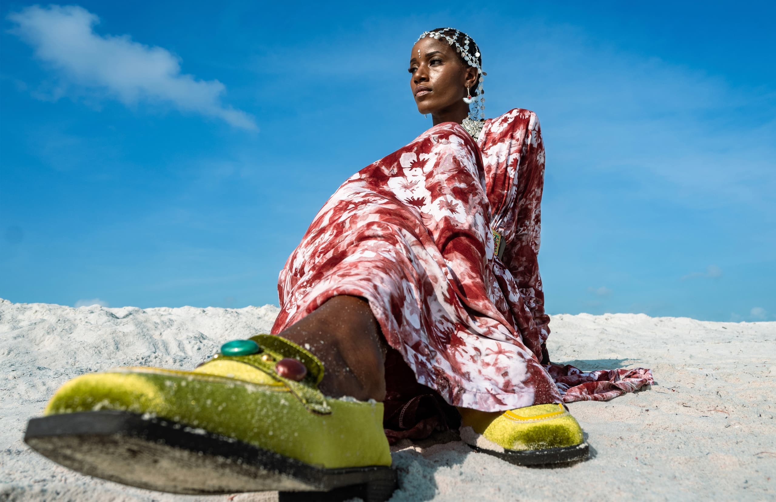 Woman in patterned dress and green shoes, sitting on a beach against a blue sky.