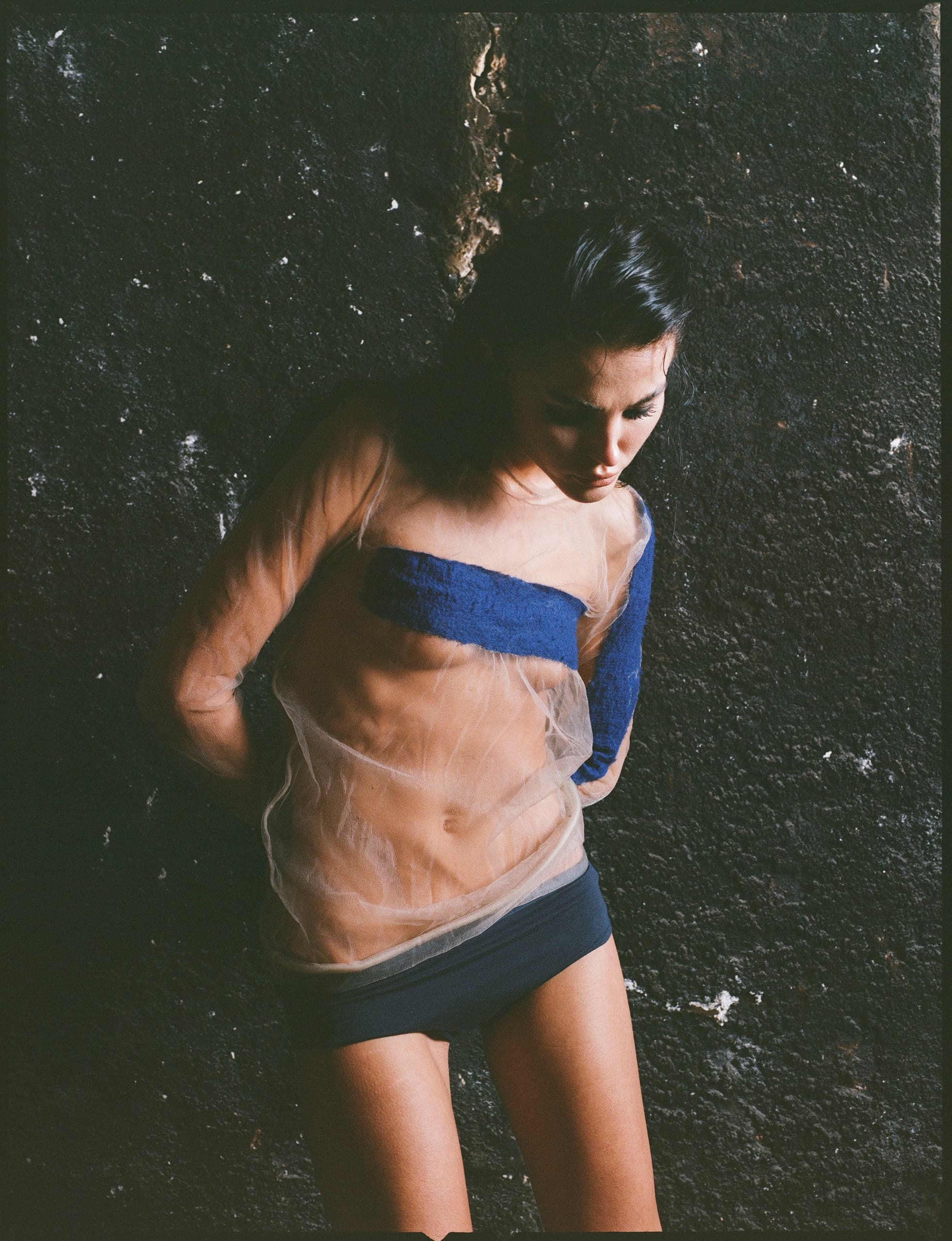 Artistic portrait of a person in sheer top against textured dark wall.