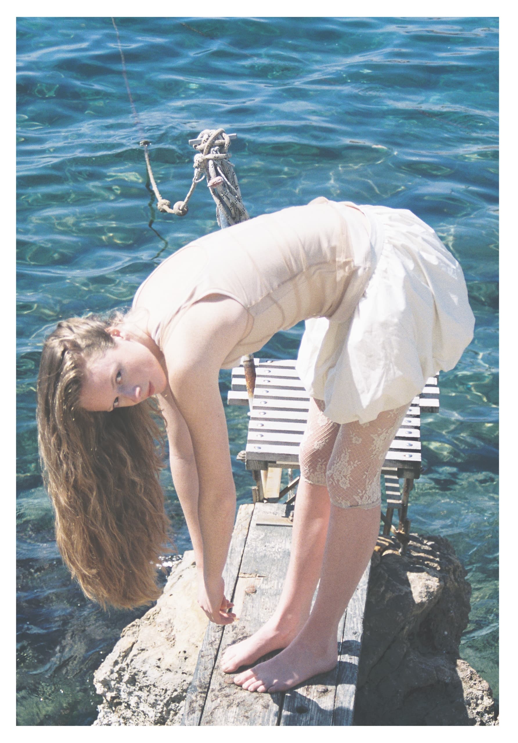 Woman bending on pier by the sea, wearing white dress and lace leggings.