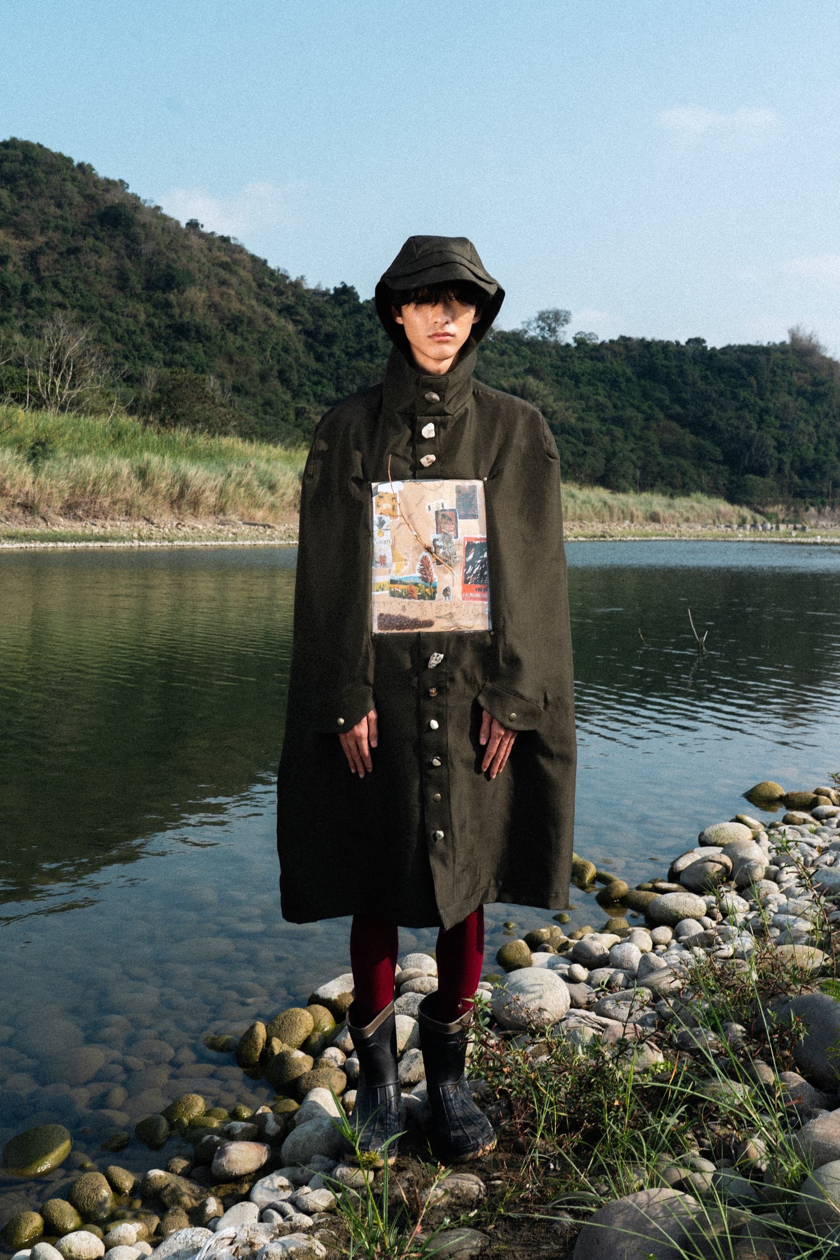 Person in dark coat by a riverside with hills in the background.