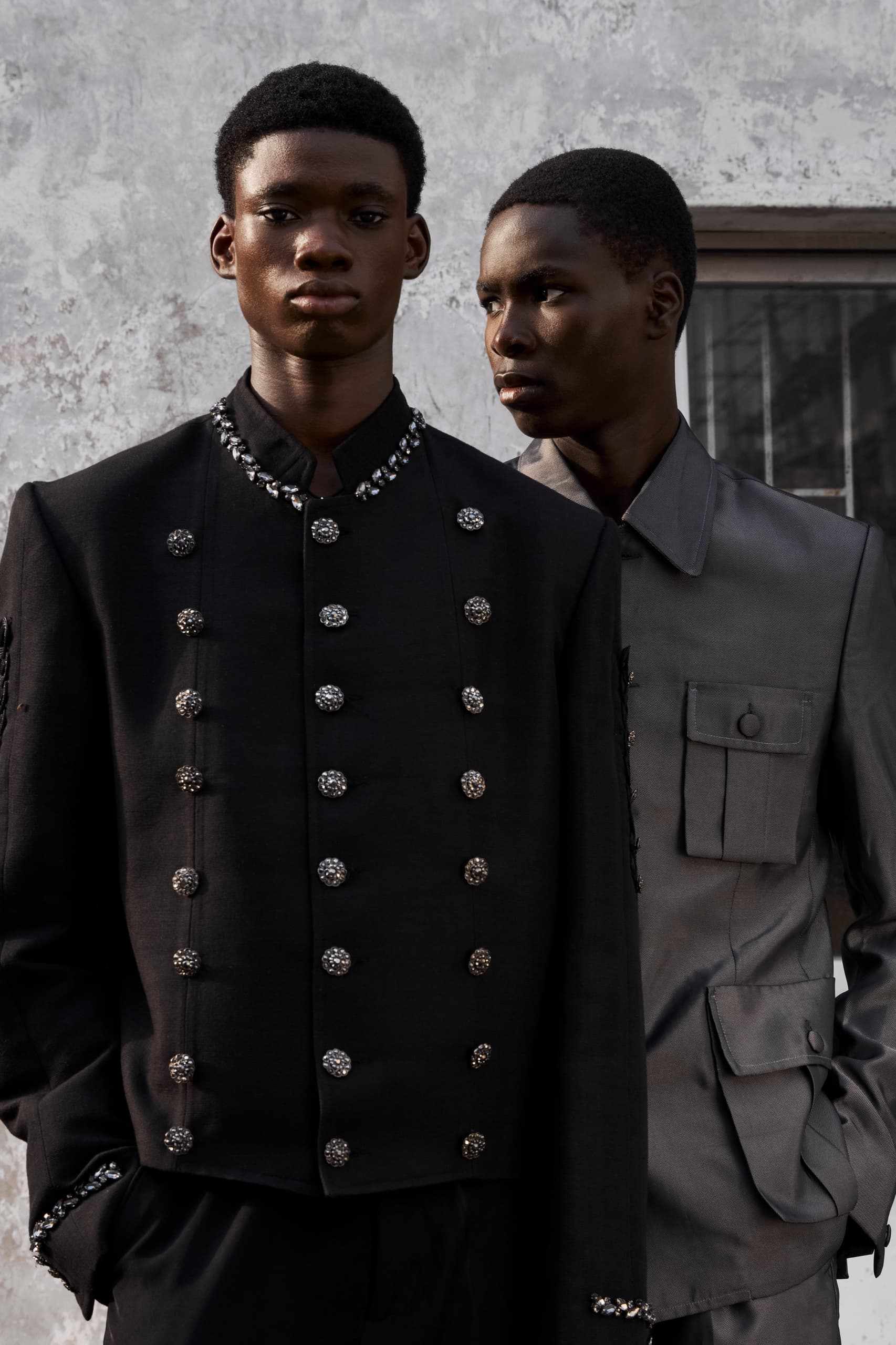 Two models in elegant embellished jackets, contrasting against a textured wall.