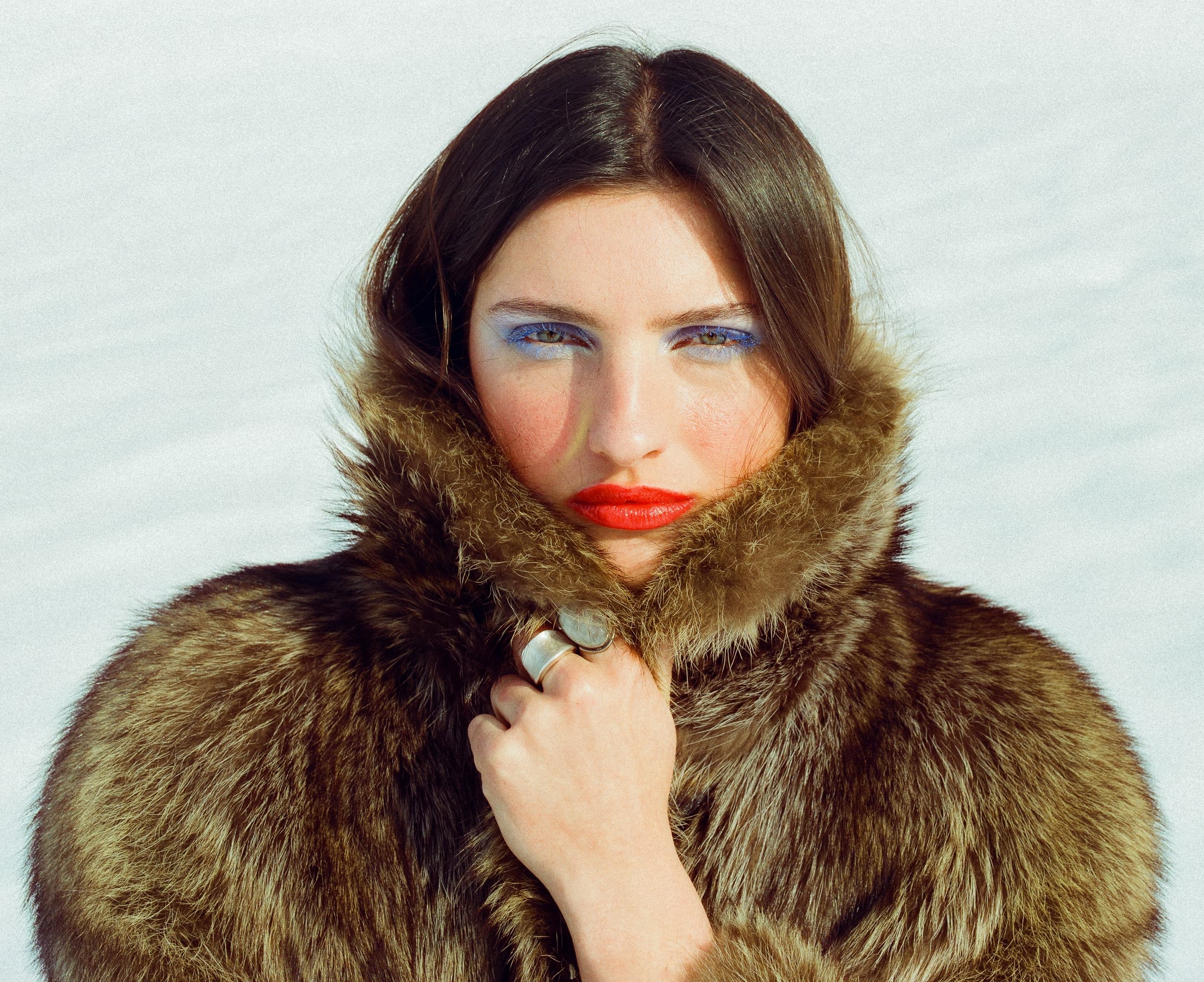 Woman in fur coat with bold makeup poses against snowy backdrop.