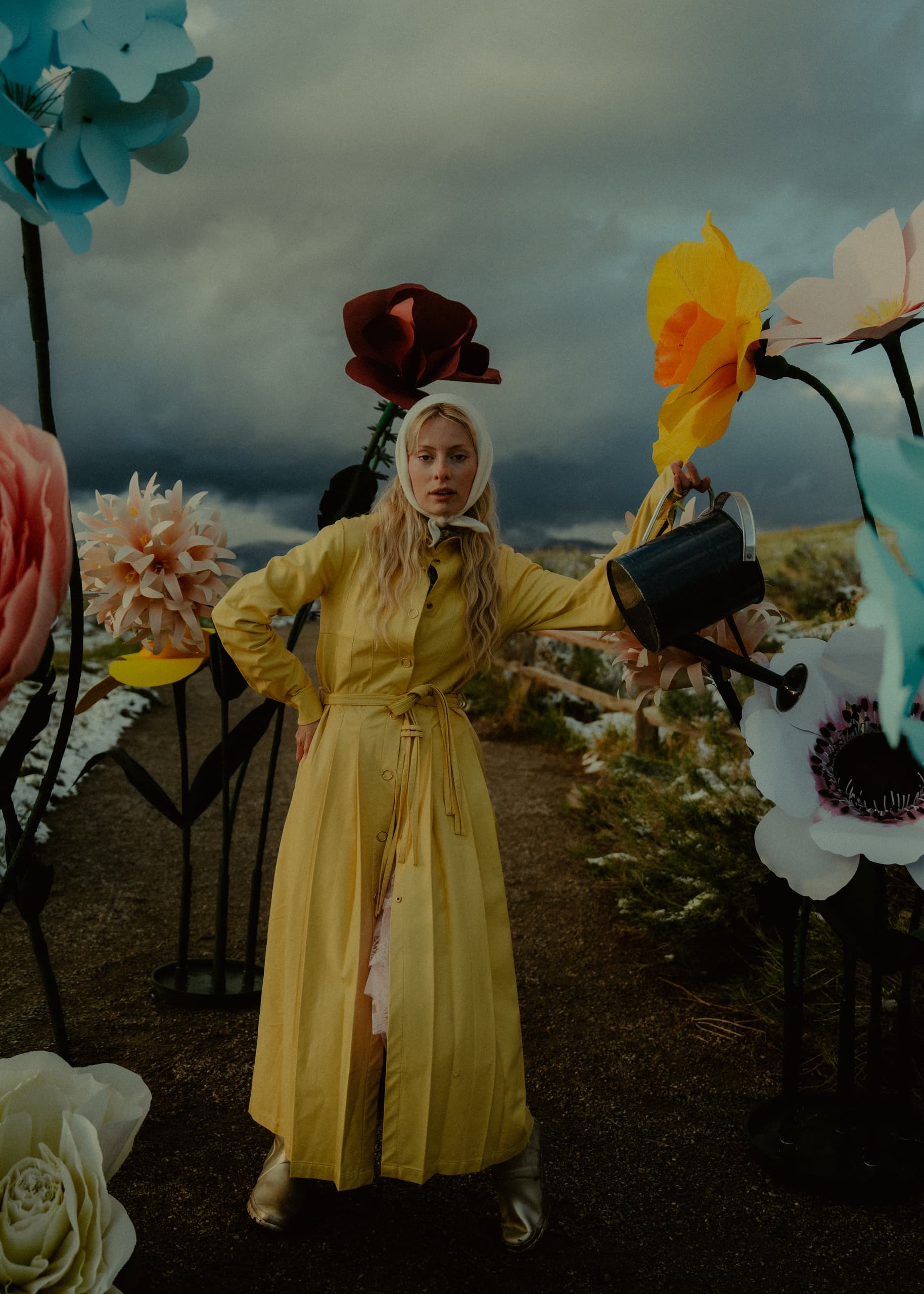 Woman in yellow coat with giant flowers, holding watering can in dreamy garden.