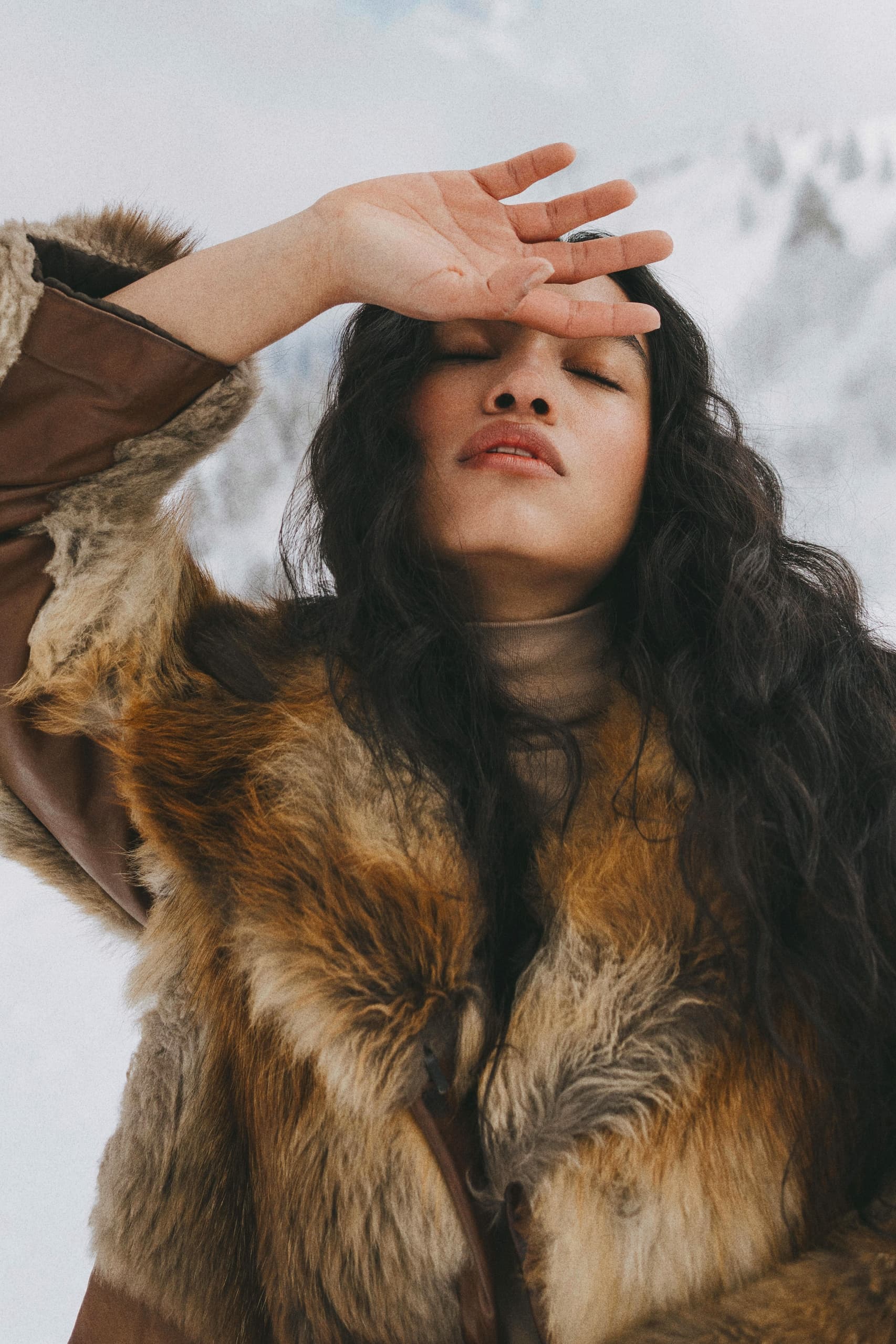 Woman in fur coat poses with hand shielding face in snowy mountain backdrop.