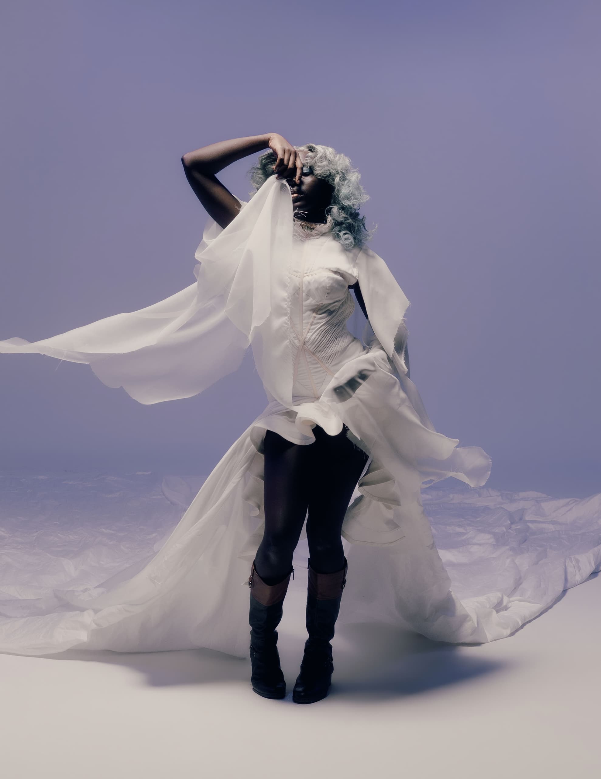 Elegant figure poses in flowing white dress with lavender backdrop.