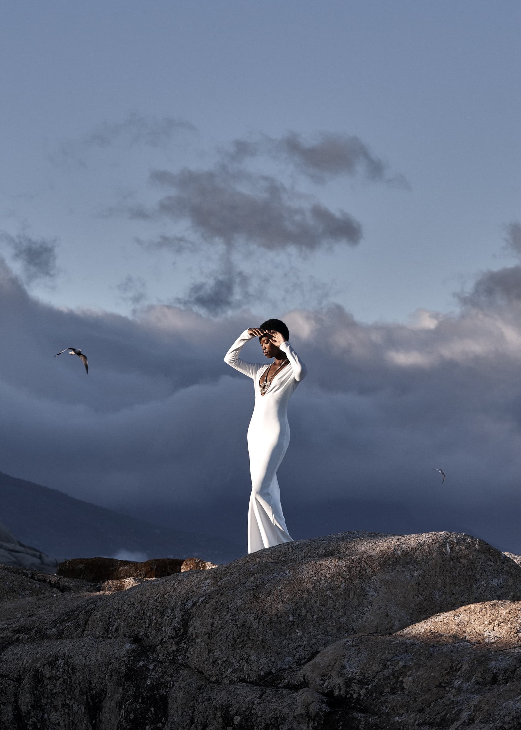 Person in elegant white dress poses on rocky hill under cloudy sky.