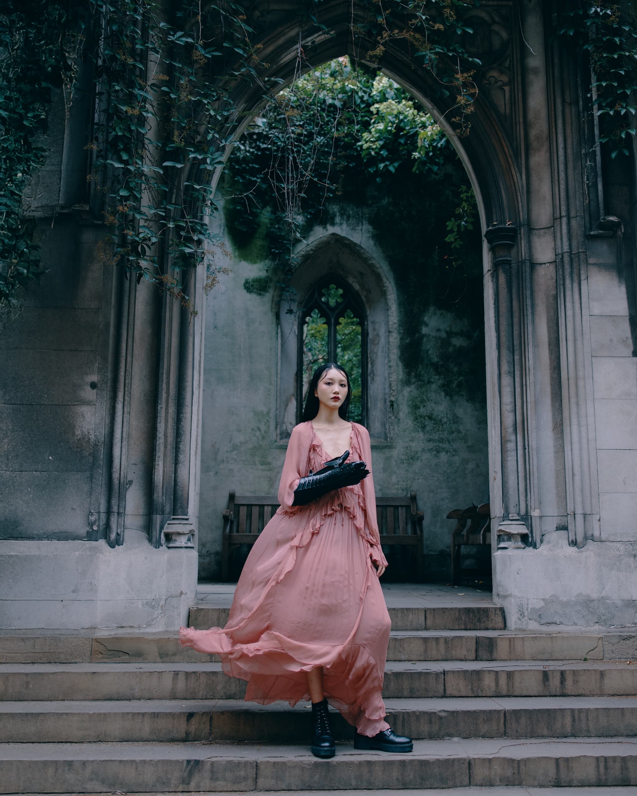 Woman in flowing pink gown on stone steps beneath ivy-covered Gothic archway.