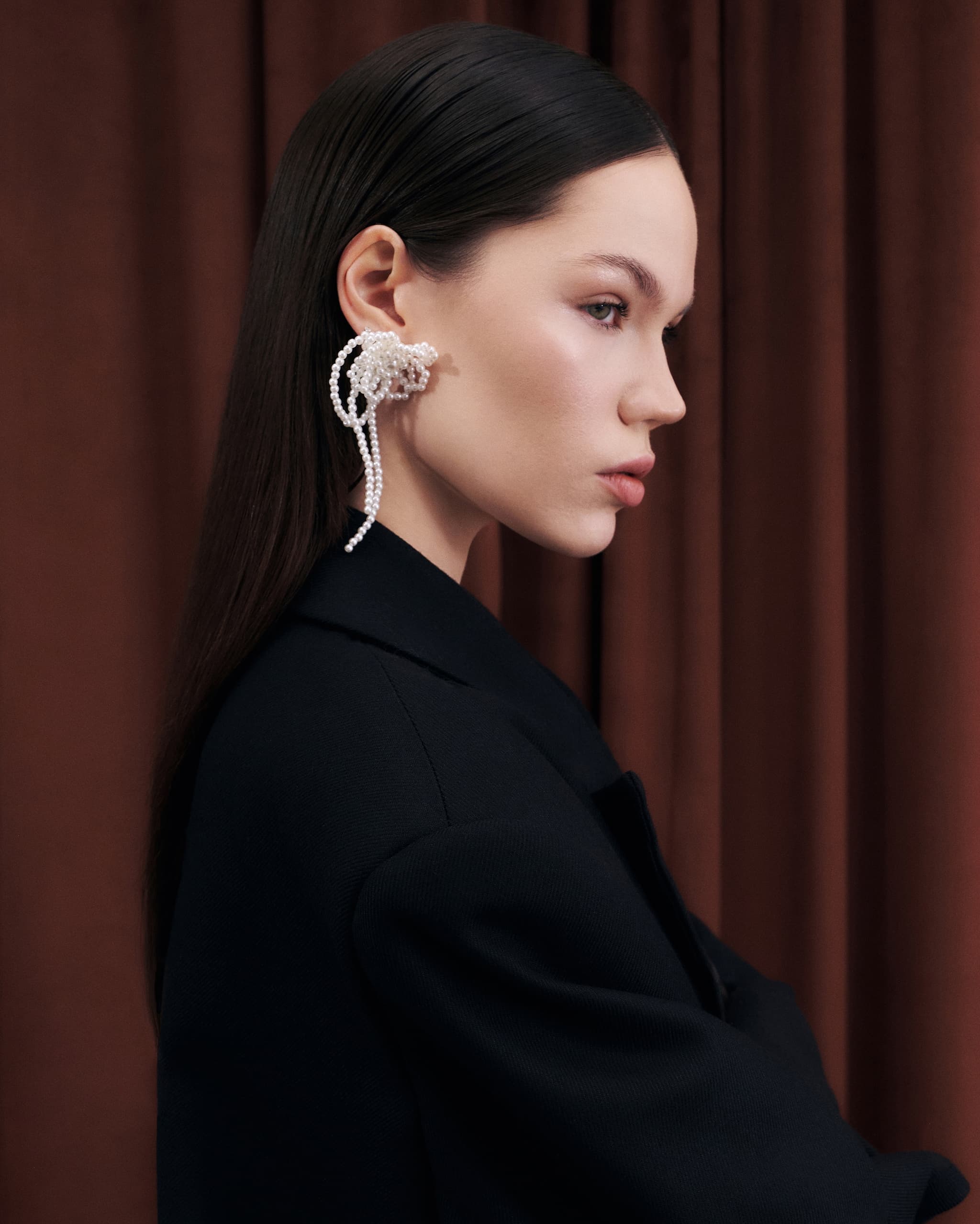 Elegant woman wearing statement pearl earring, side profile against brown drapery.