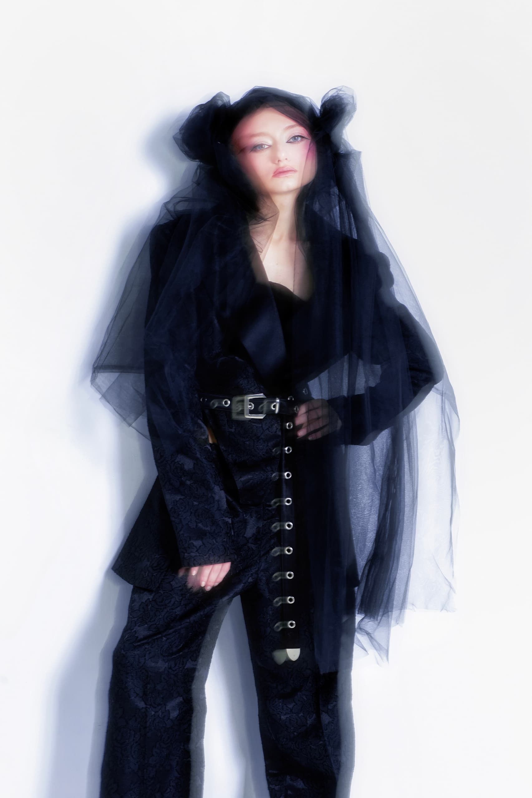 Fashion model in a dark, ethereal outfit with a sheer veil, set against a white background.