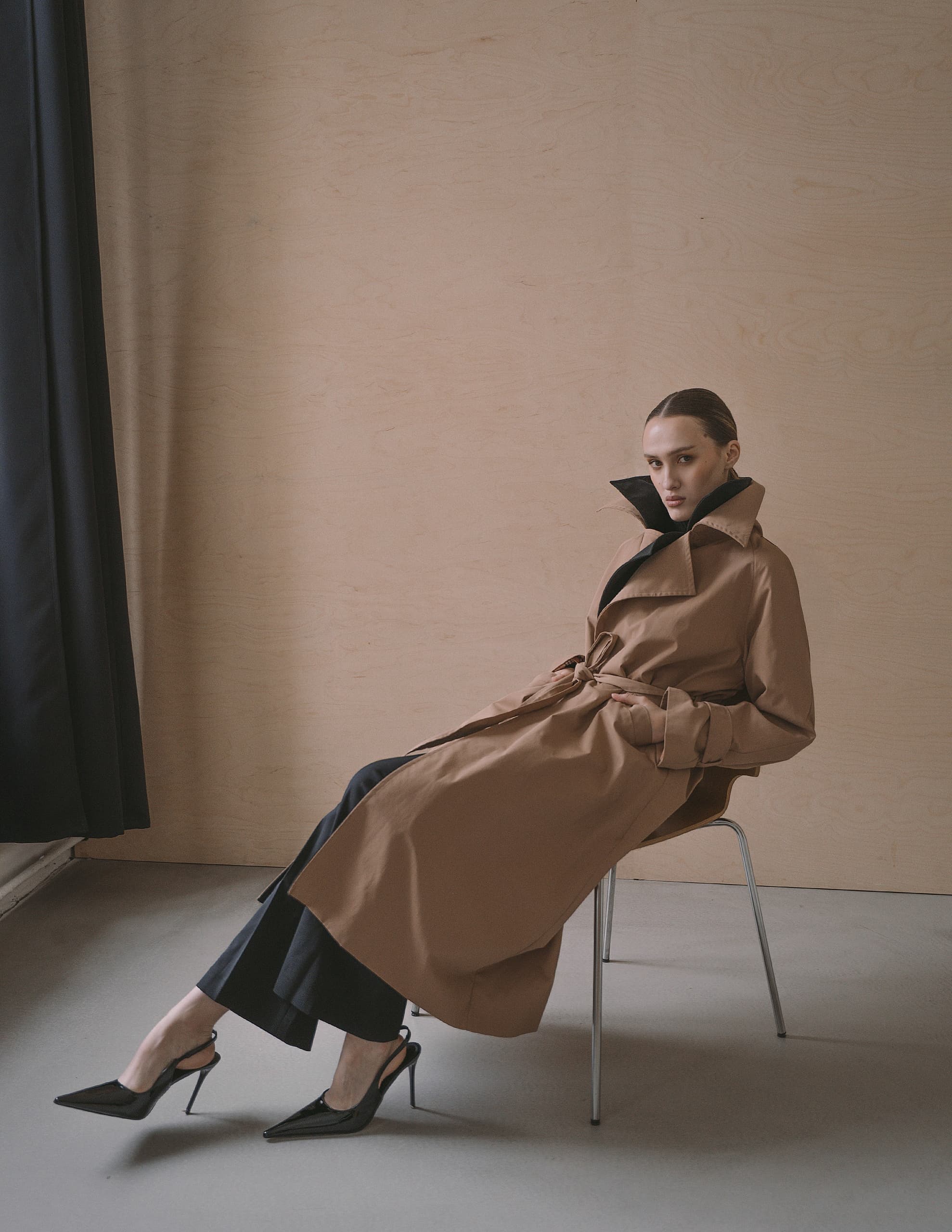 Elegant woman in brown trench coat seated, wearing black heels.