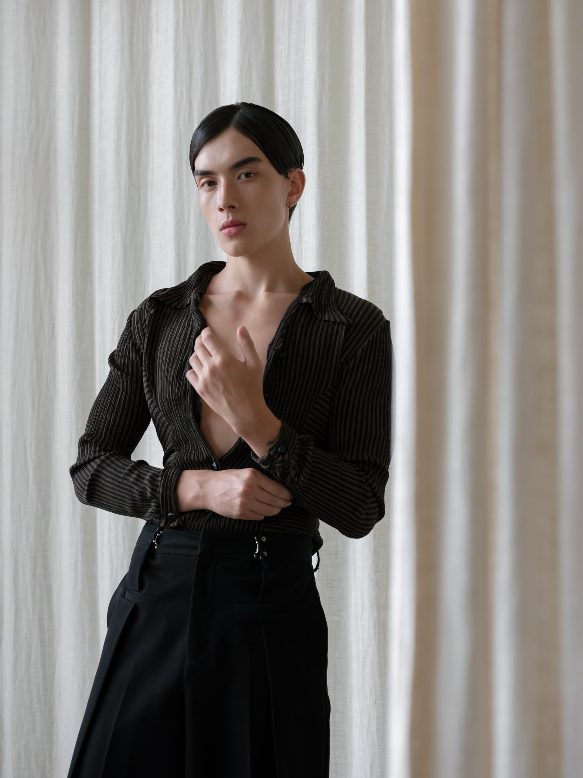 Person in black striped shirt standing by curtains, striking a confident pose.