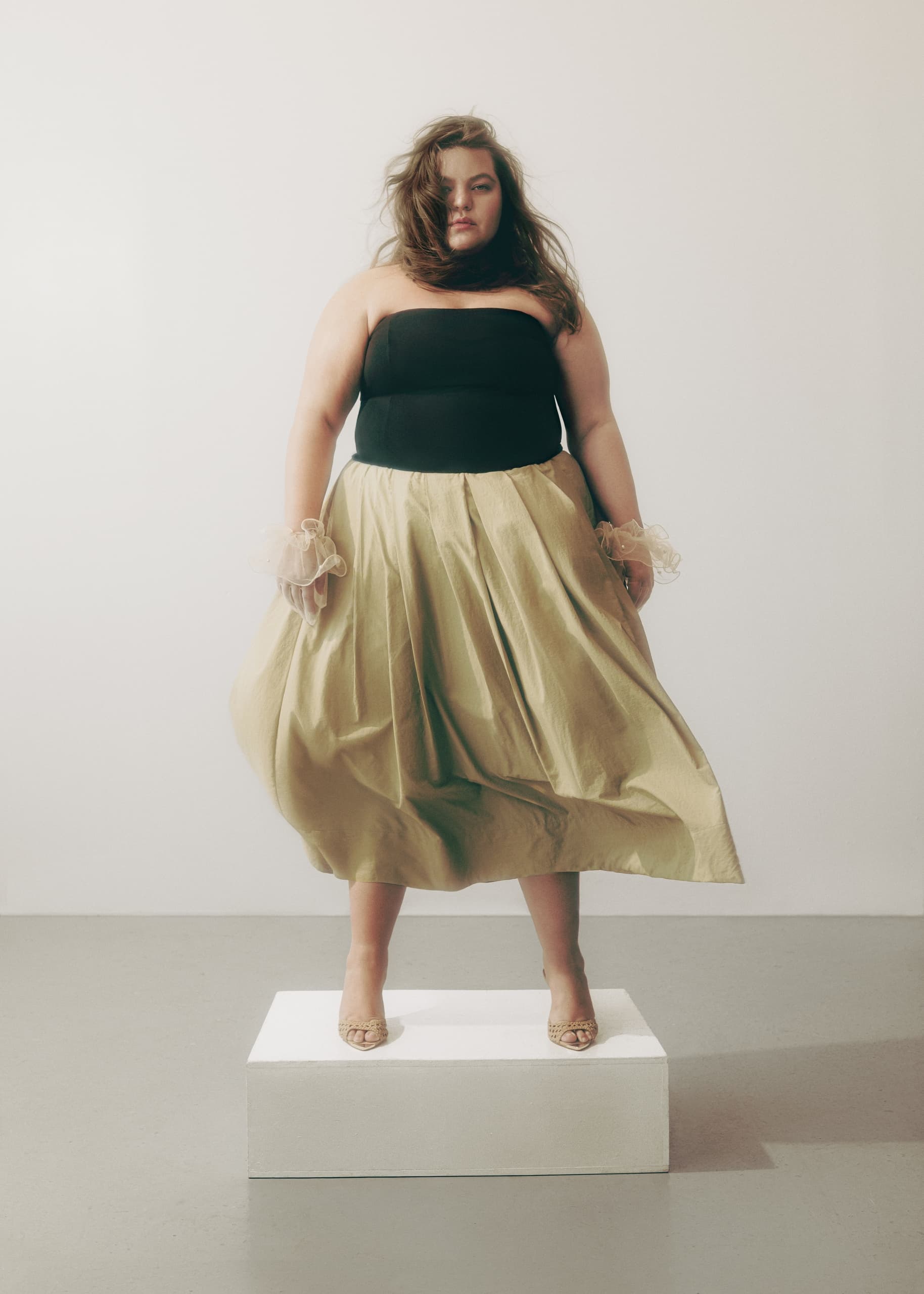Fashion portrait of a model in a black top and gold skirt on a white pedestal.