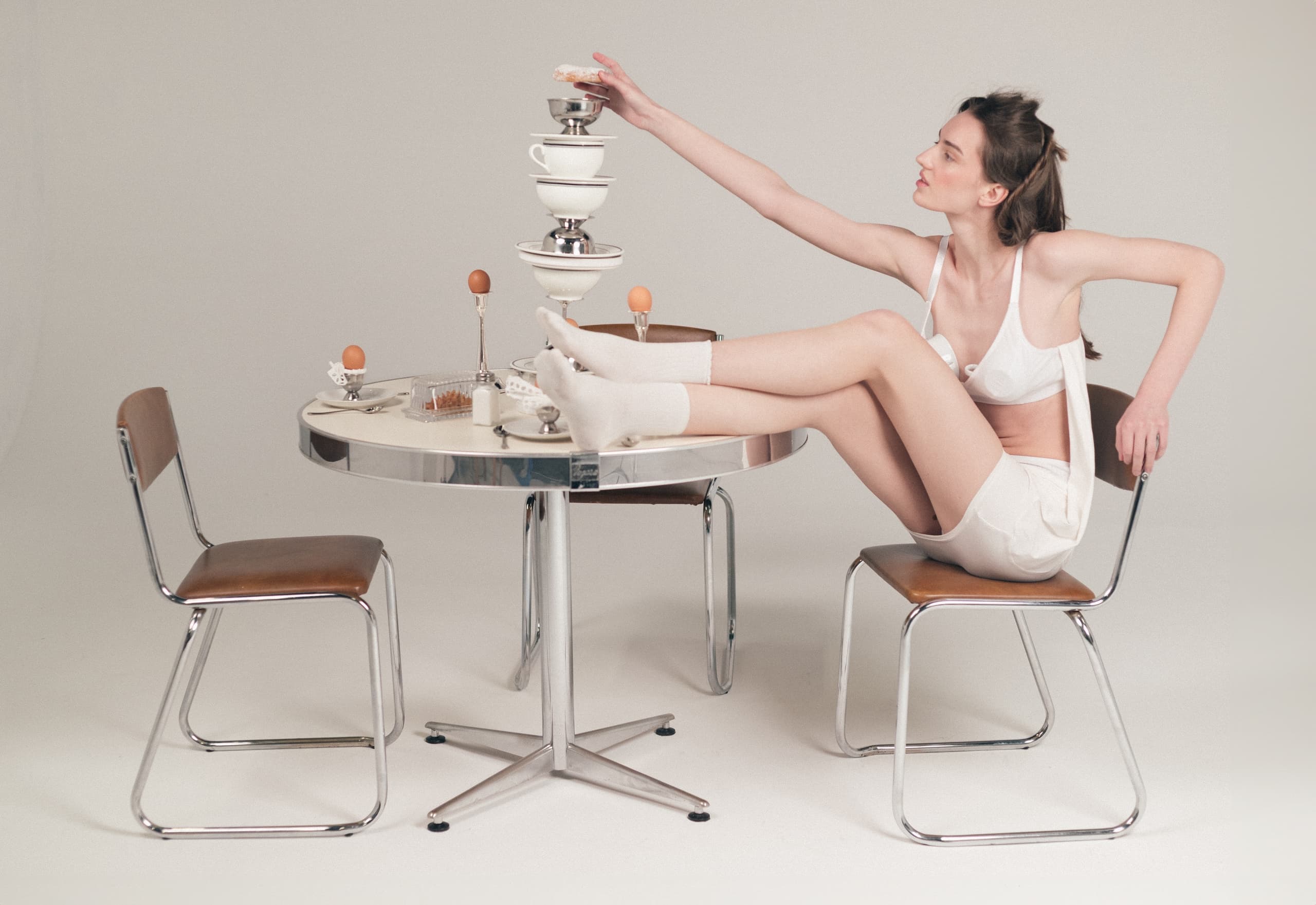 Woman in white lingerie balancing cups on a table, modern minimalist setting.