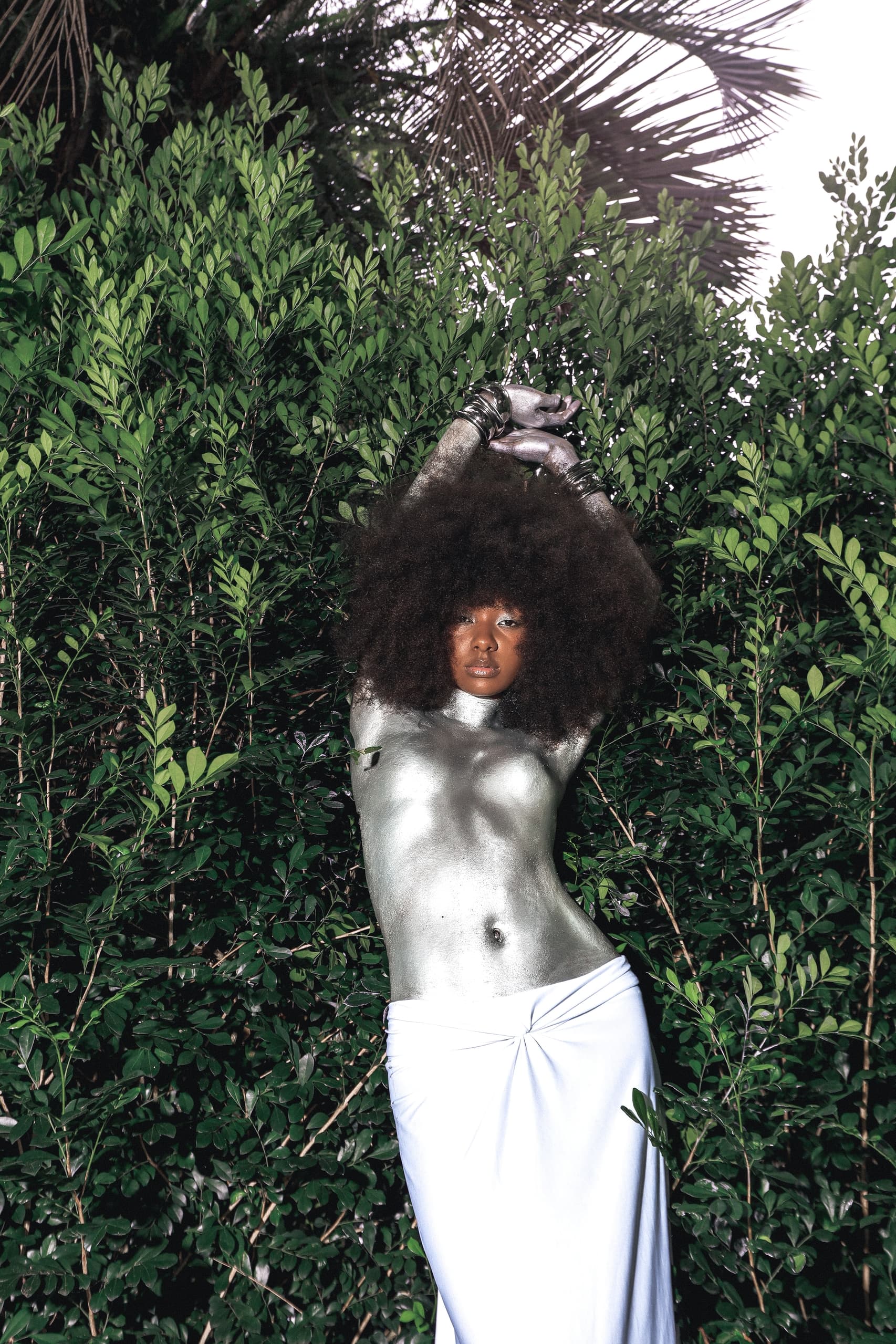 Person painted in silver posing in front of lush green foliage