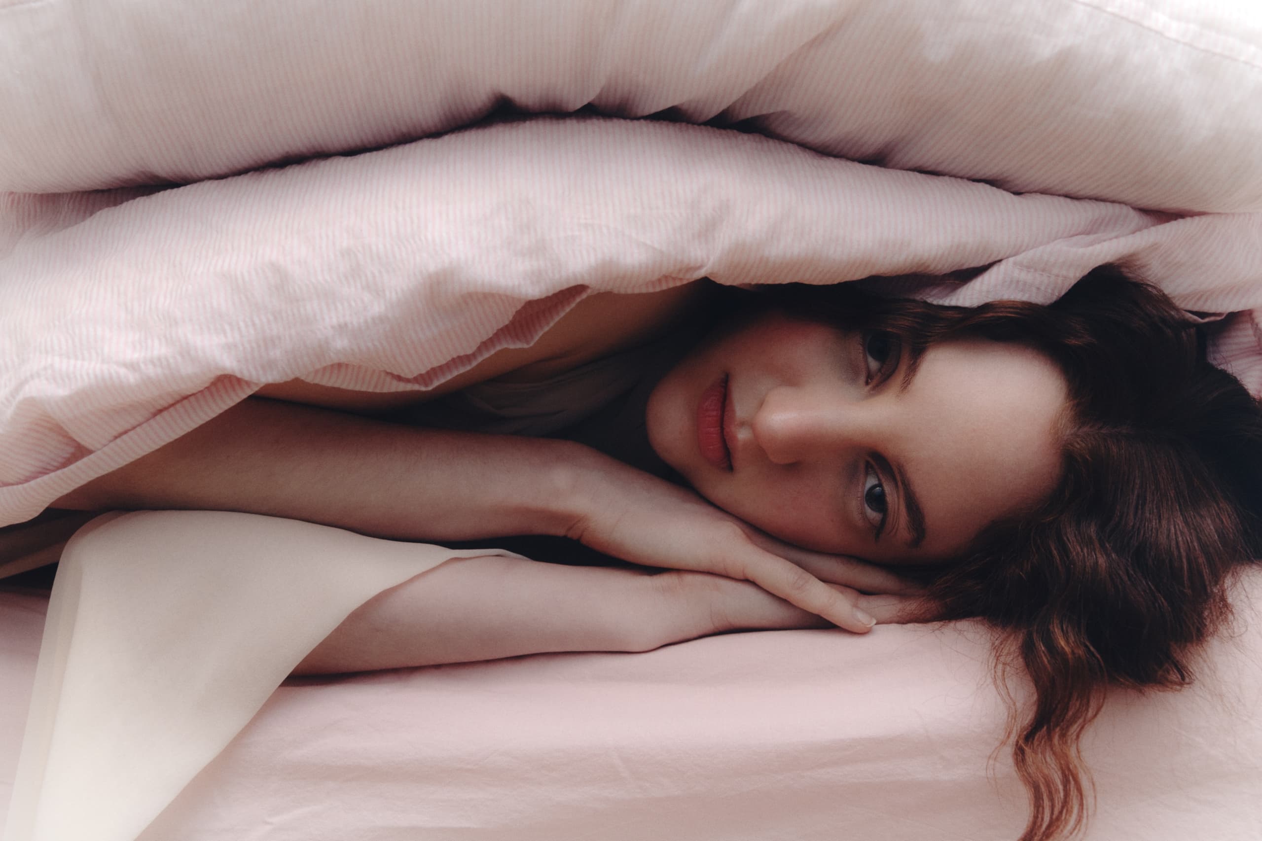 Cozy woman under pink duvet blanket, serene expression, resting head on pillow.