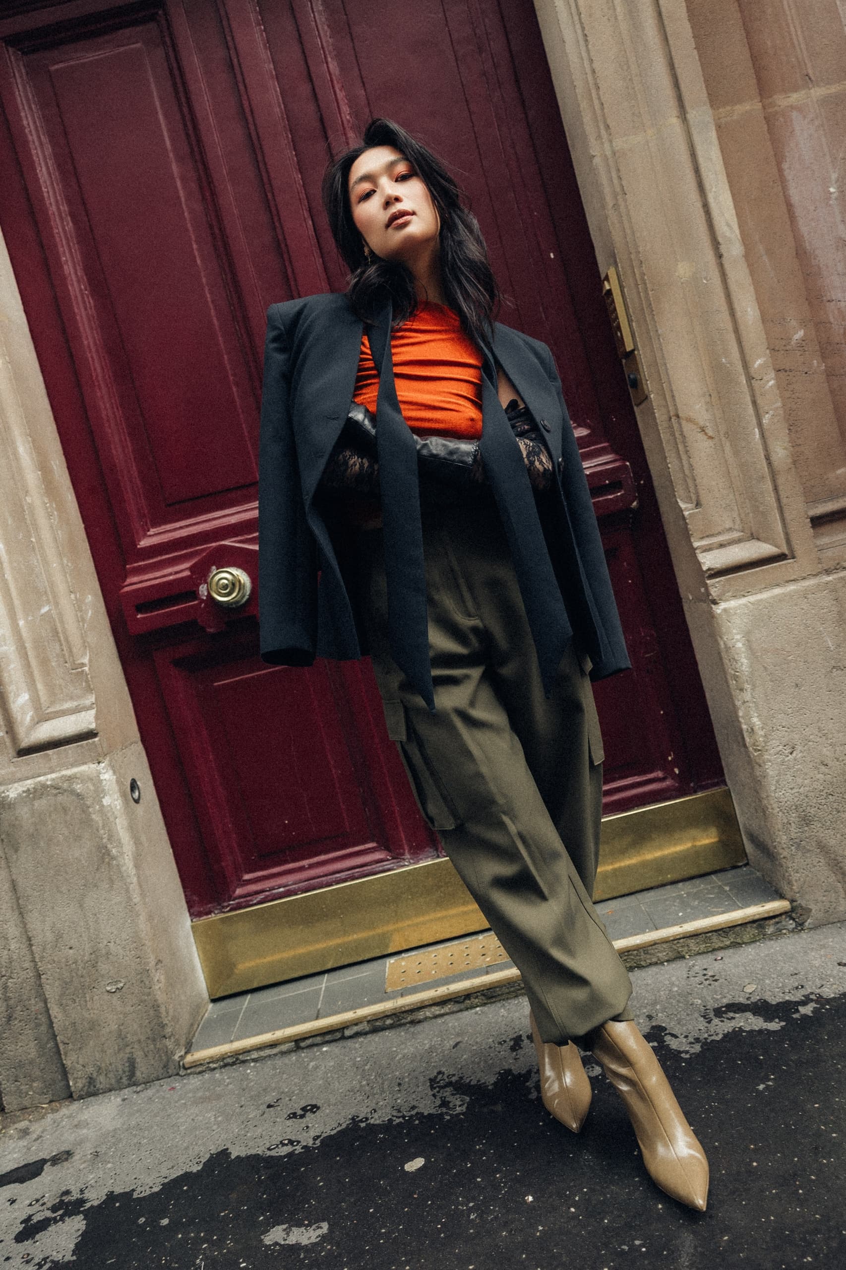 Fashionable woman in orange top and blazer poses by elegant red doorway.