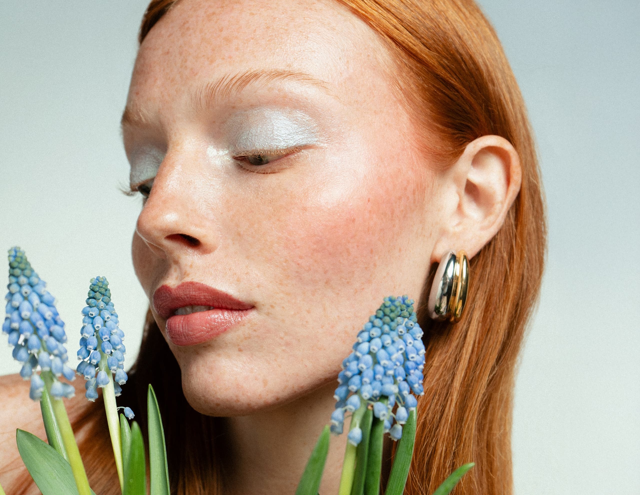 Woman with red hair and silver eye makeup near blue flowers.