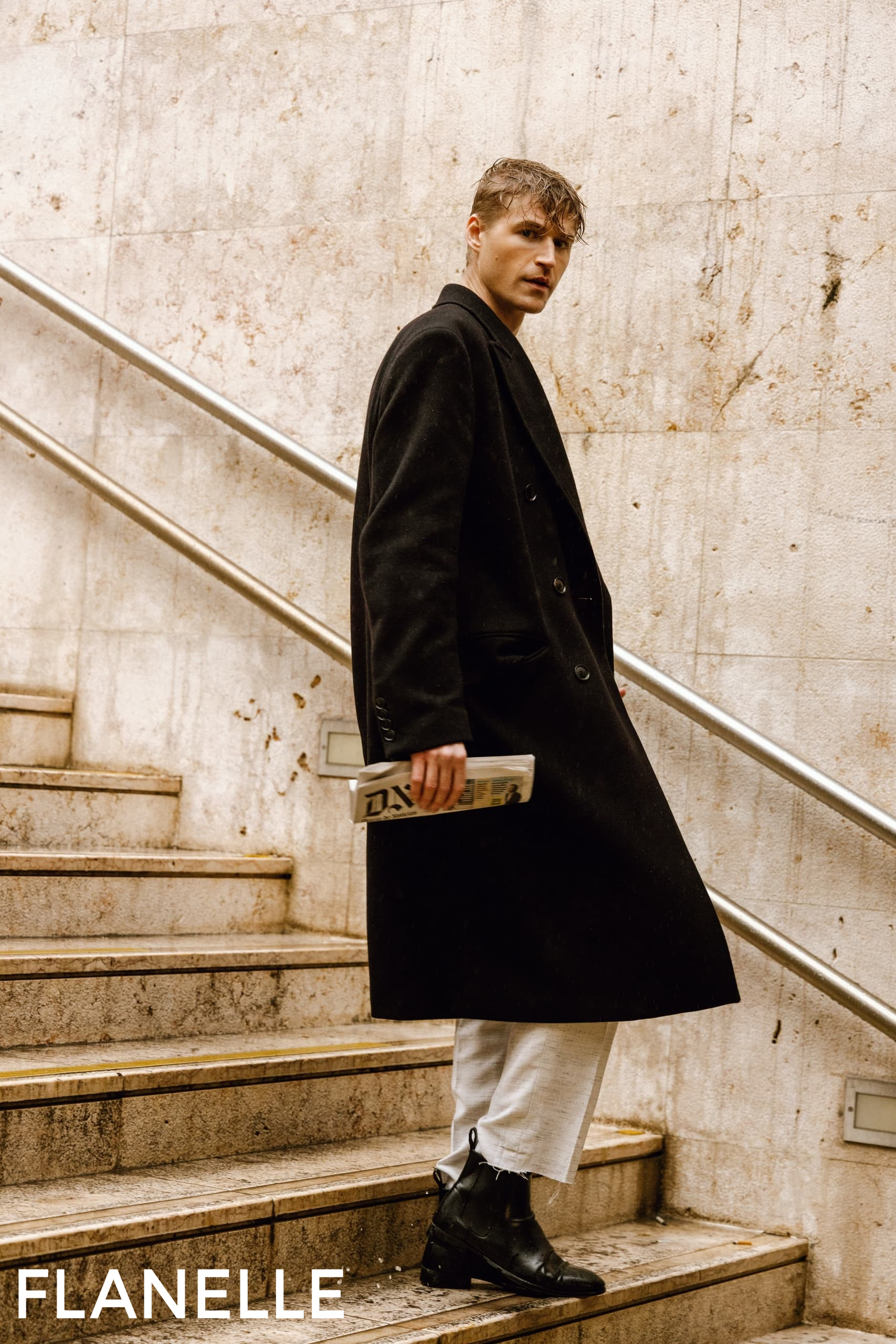 Man in stylish coat ascending stairs, holding newspaper, urban fashion scene.