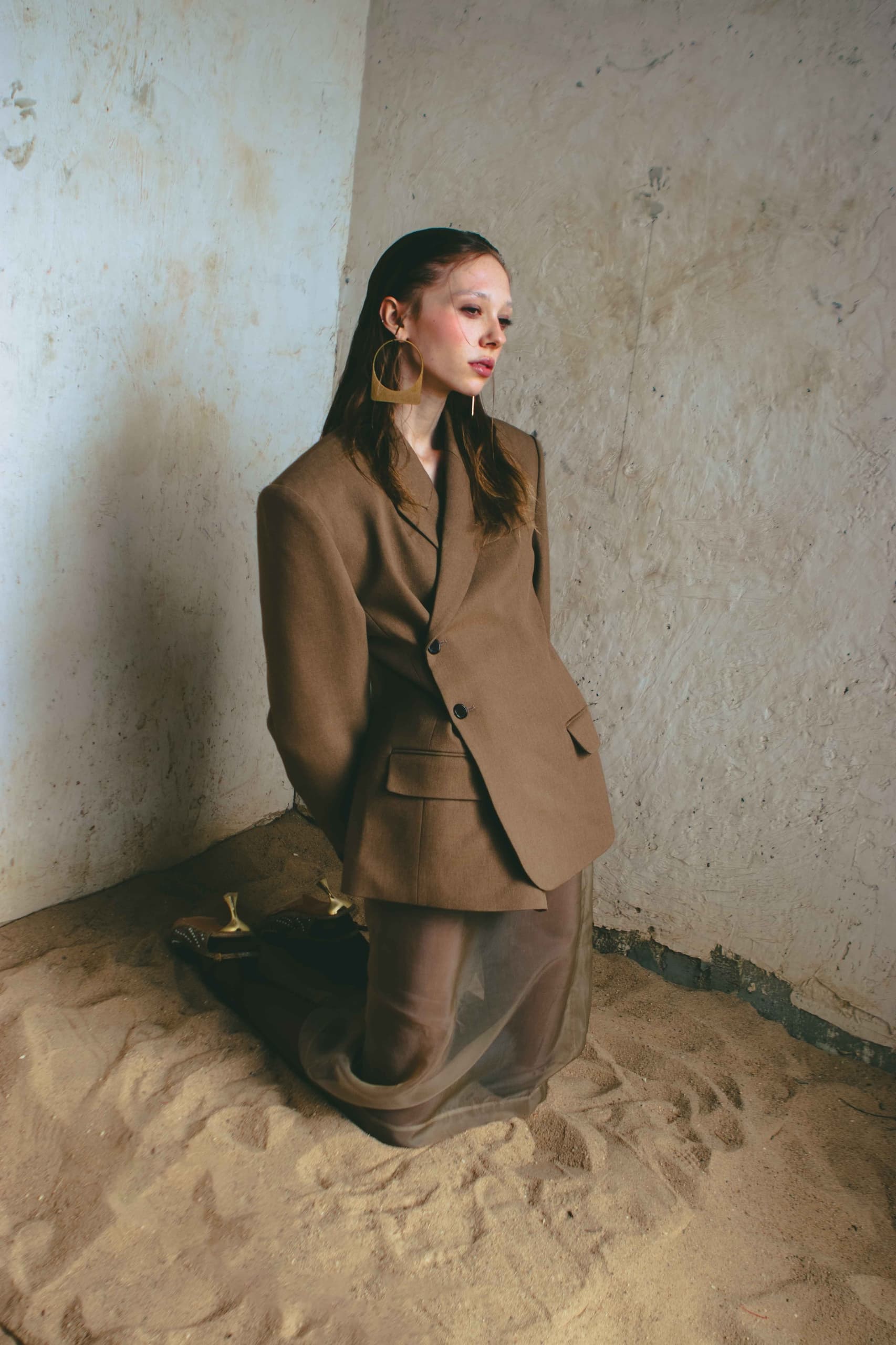 Fashion model in brown suit kneels on sandy floor near textured concrete wall.